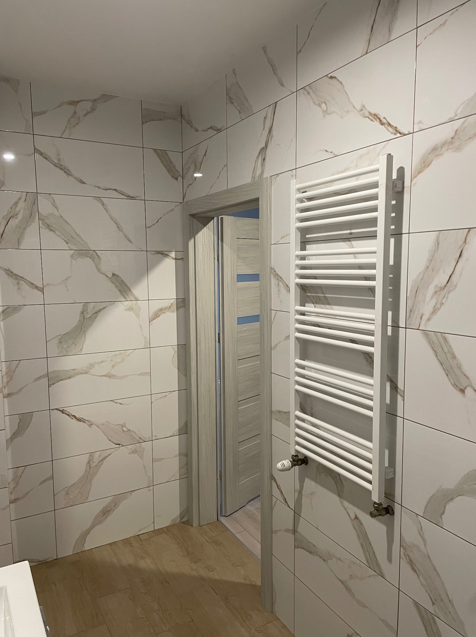 Wykończona łazienka z marmurowymi płytkami, grzejnikiem drabinkowym i drewnopodobną podłogą. Widoczne drzwi z ościeżnicą w odcieniach szarości.