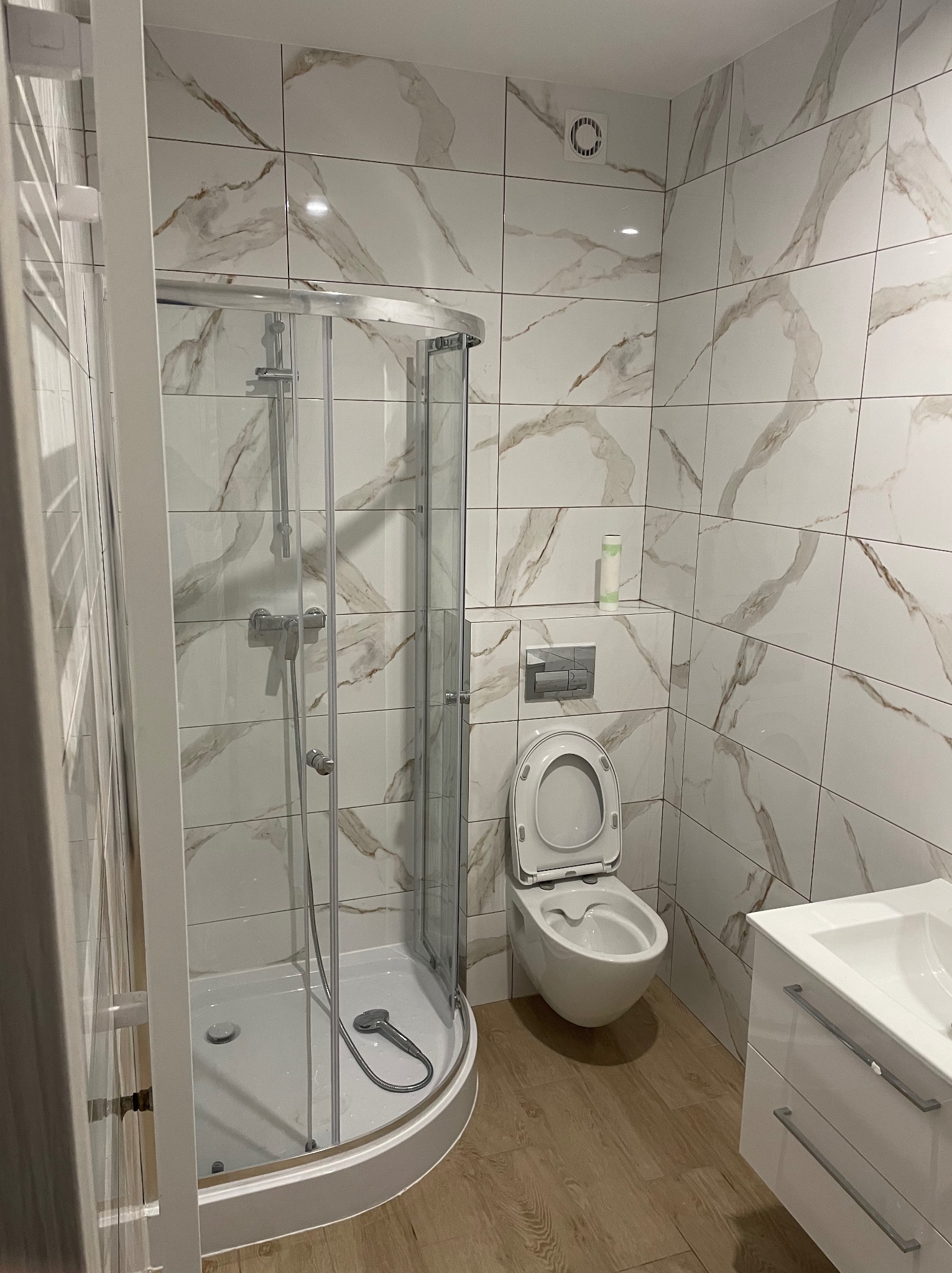 Nowoczesna łazienka z kabiną prysznicową, toaletą i umywalką w zabudowie. Białe płytki z marmurkowym wzorem, drewnopodobna podłoga, chromowana armatura.