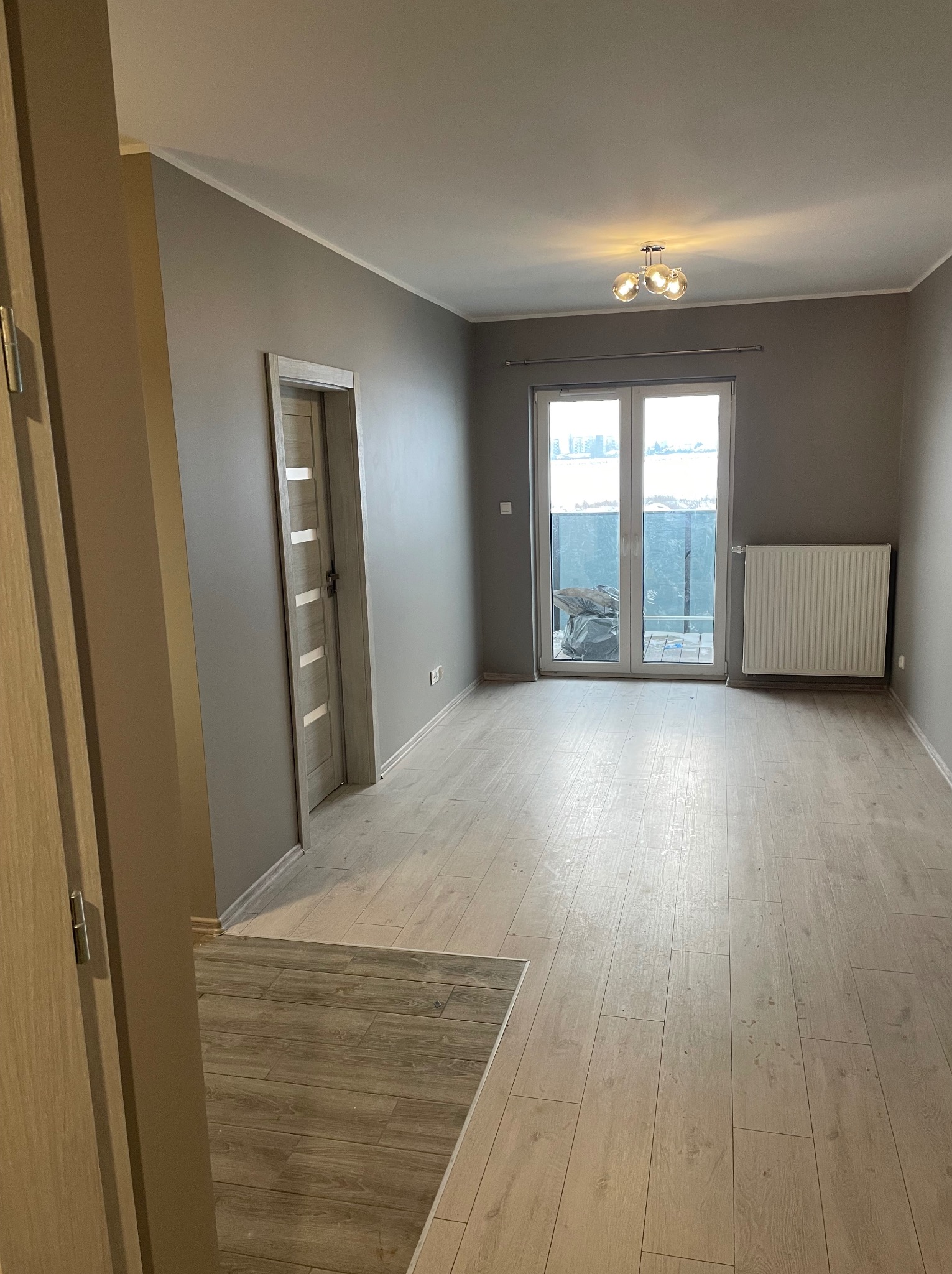 Szare, puste wnętrze po remoncie z panelami podłogowymi, grzejnikiem i drzwiami balkonowymi. Widoczny fragment progu z innym wzorem paneli.