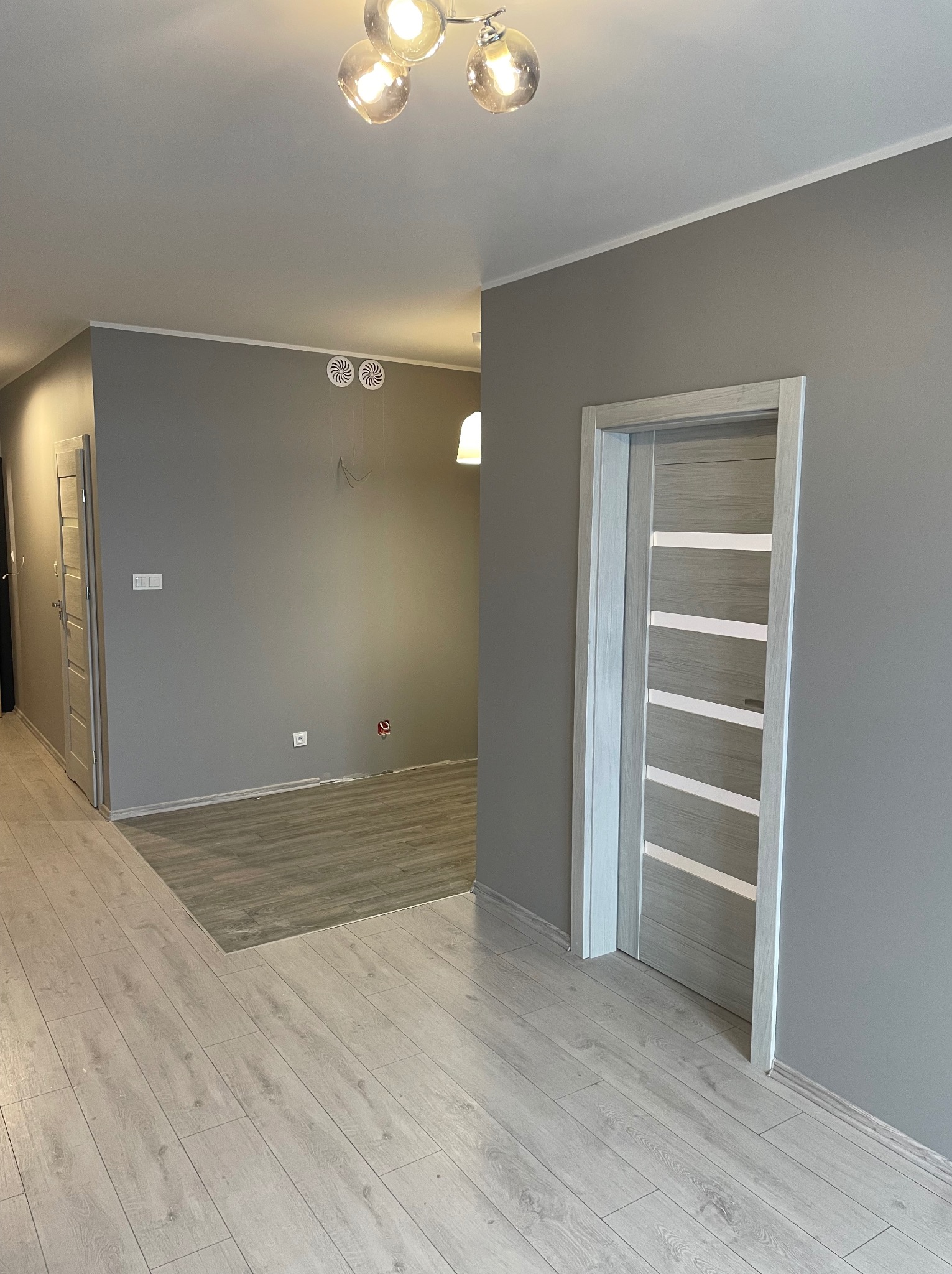 Szare wnętrze po remoncie: świeżo pomalowane ściany, jasna podłoga, nowoczesne drzwi z poziomymi paskami i designerska lampa sufitowa.