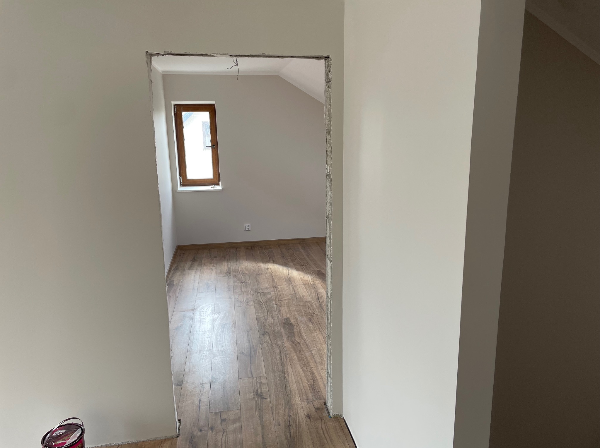 Wnętrze pokoju na poddaszu z drewnianą podłogą, widoczne przez surowy otwór drzwiowy. Ściany pomalowane na jasny kolor, w tle okno w drewnianej ramie.