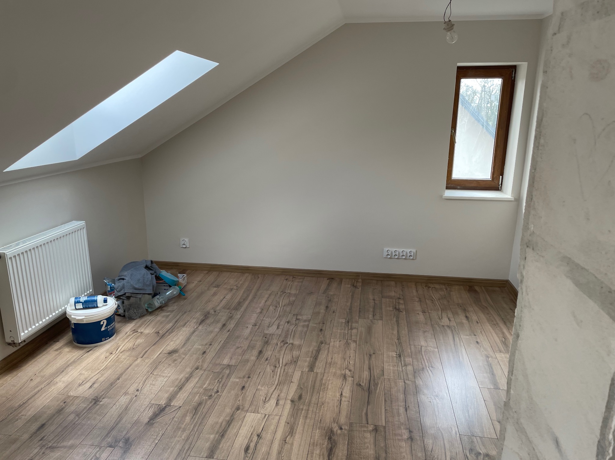 Poddasze po malowaniu ścian na neutralny odcień, z drewnianą podłogą, oknem dachowym i pionowym. Widoczne akcesoria malarskie i grzejnik.