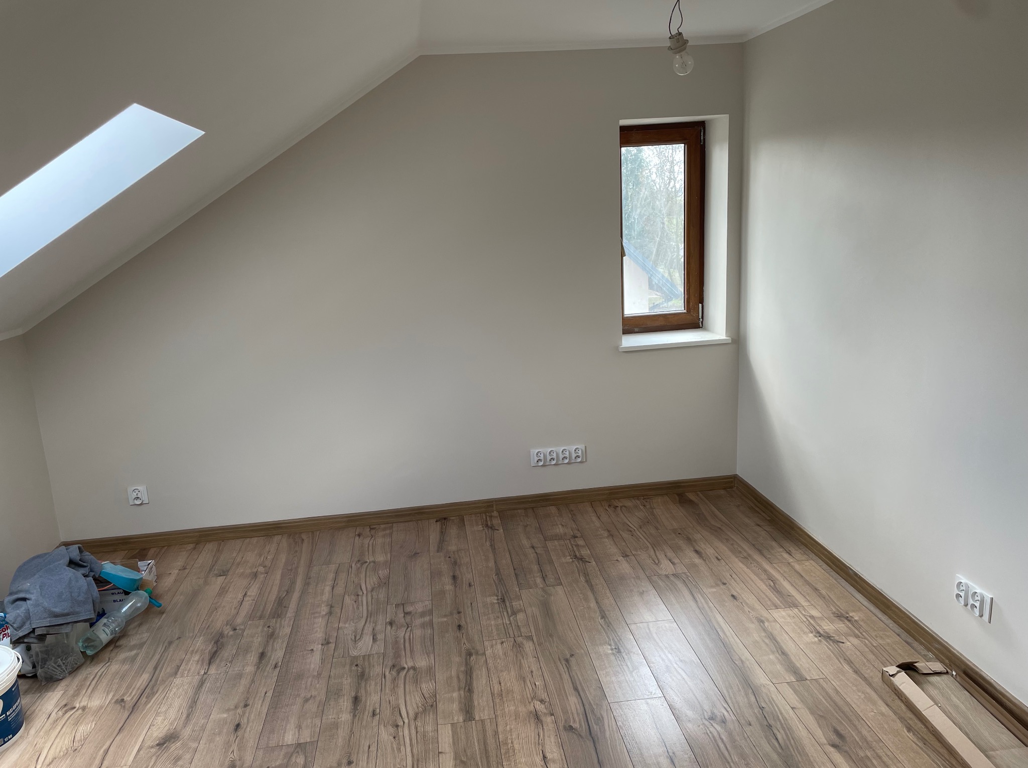 Poddasze po malowaniu w Gostyninie. Widoczne okno dachowe, drewniana podłoga i świeżo pomalowane ściany na biało. Częściowo widoczne materiały malarskie.