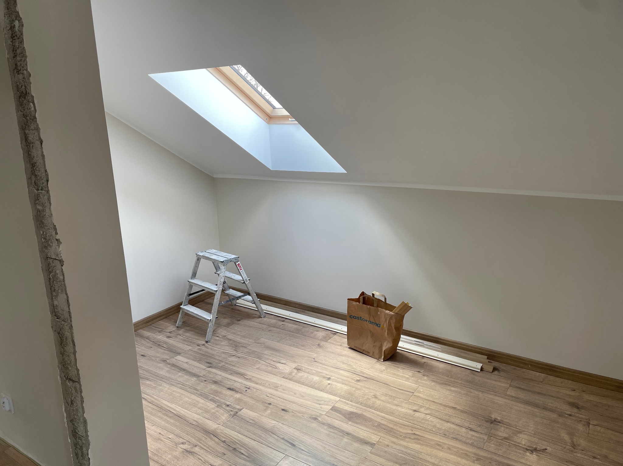 Poddasze z oknem dachowym, drewnianą podłogą, drabinką i torbą Castorama. Widoczny fragment ściany w stanie surowym. Etap prac wykończeniowych.