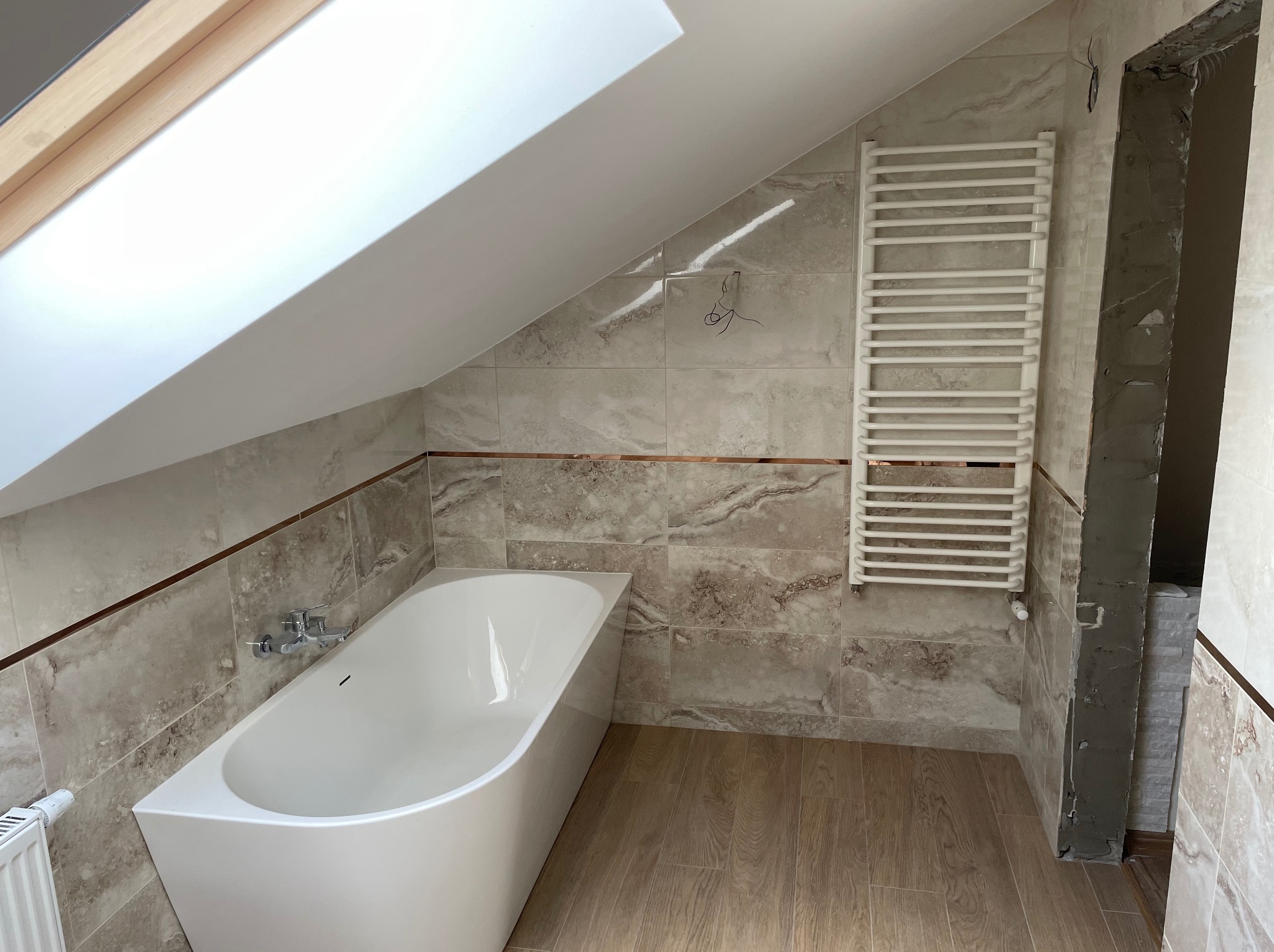 Wykończona łazienka na poddaszu z wanną, grzejnikiem drabinkowym i marmurowymi płytkami. Widoczne okno dachowe i fragment niedokończonego przejścia.