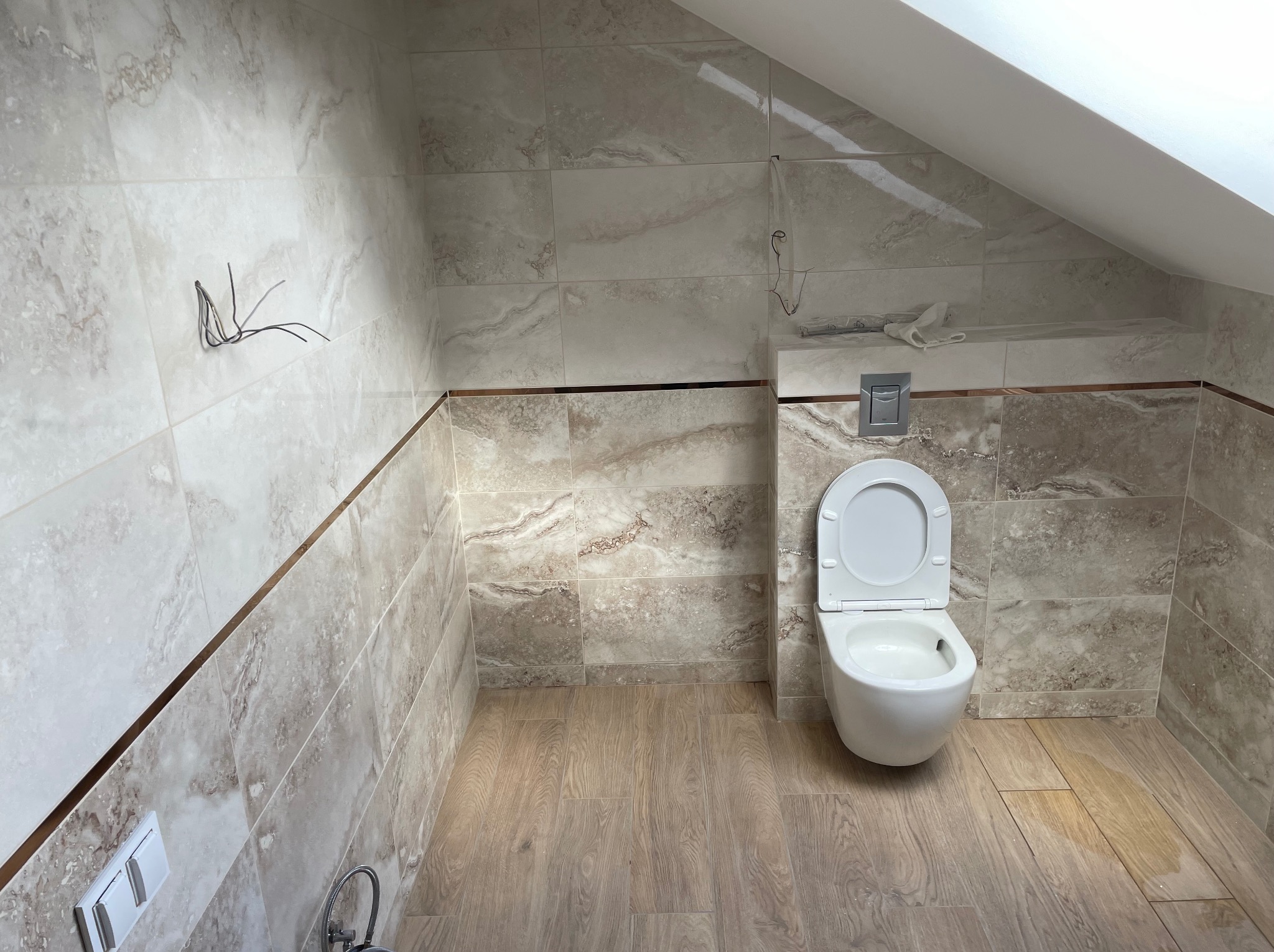 Wykończona łazienka na poddaszu ze skośnym sufitem, z beżowymi płytkami imitującymi marmur i drewnopodobną podłogą. Wisząca toaleta z otwartą klapą.