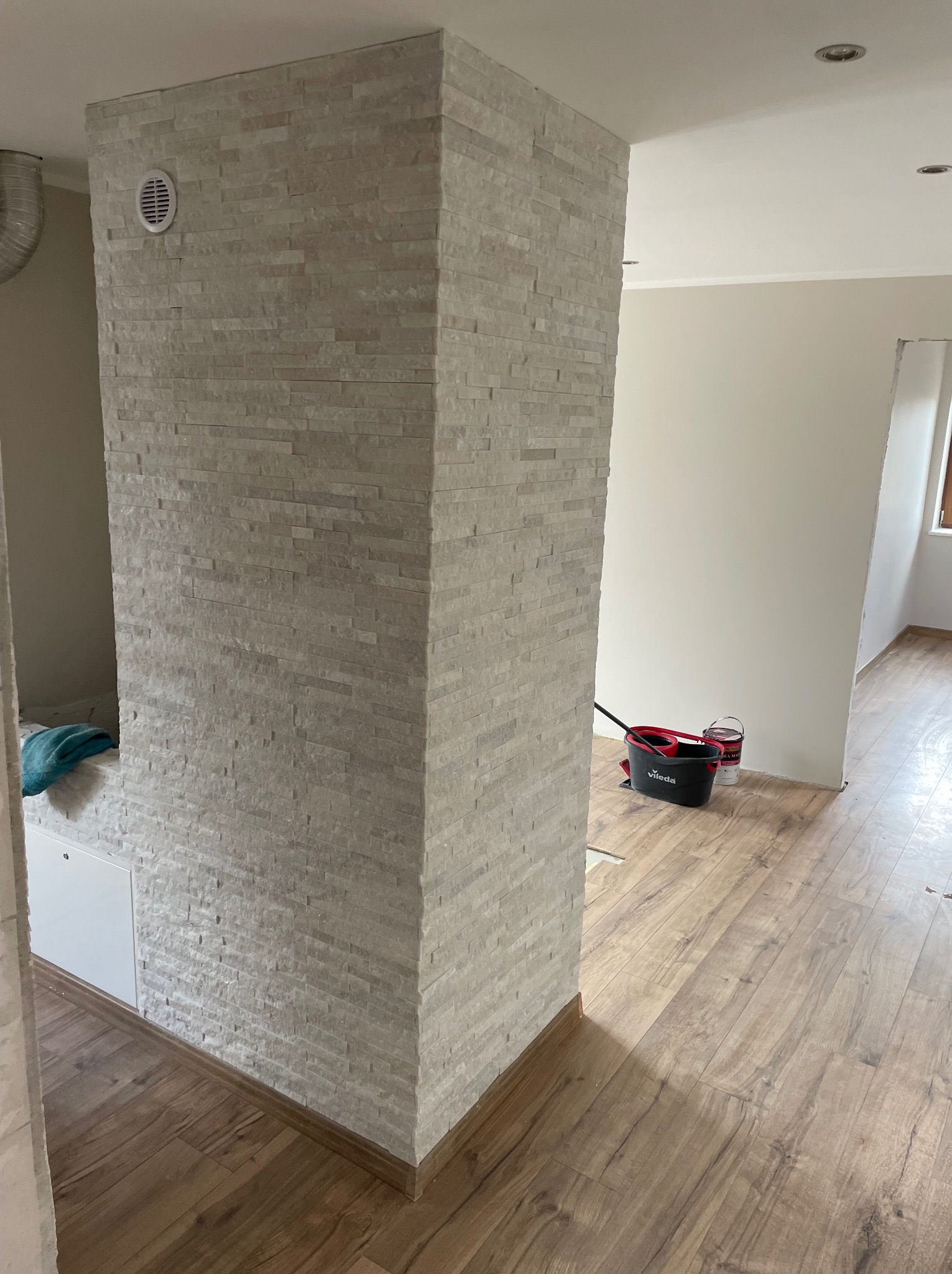 Wykończony filar z dekoracyjnej, jasnej cegły w nowoczesnym wnętrzu. Widoczna drewniana podłoga i akcesoria do sprzątania. Element dekoracyjny z wentylacją.