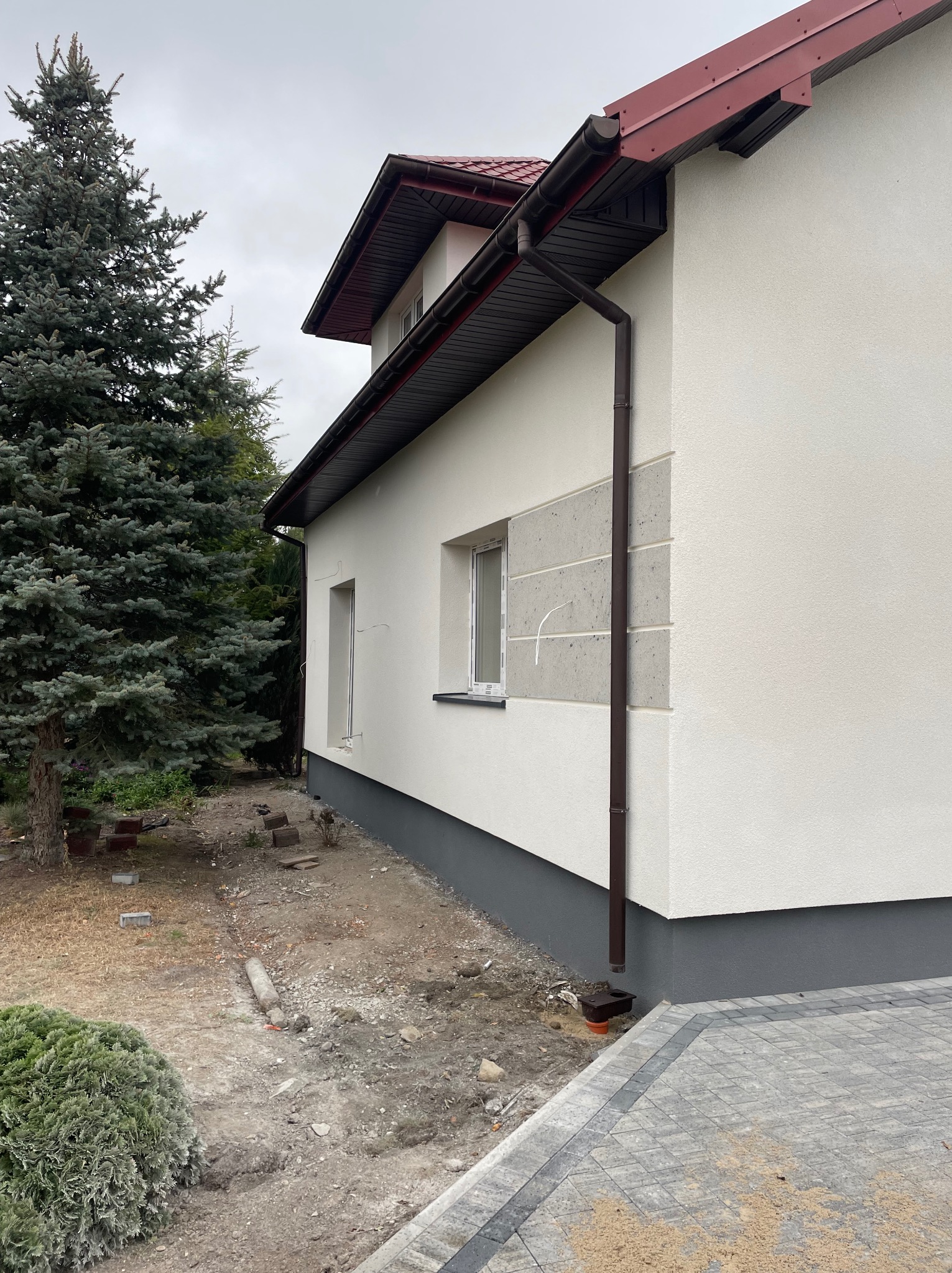 Boczny widok domu z nową, białą elewacją, brązowymi rynnami i szarą podmurówką. Fragment posesji z kostką brukową i zielenią. Pochmurne niebo.