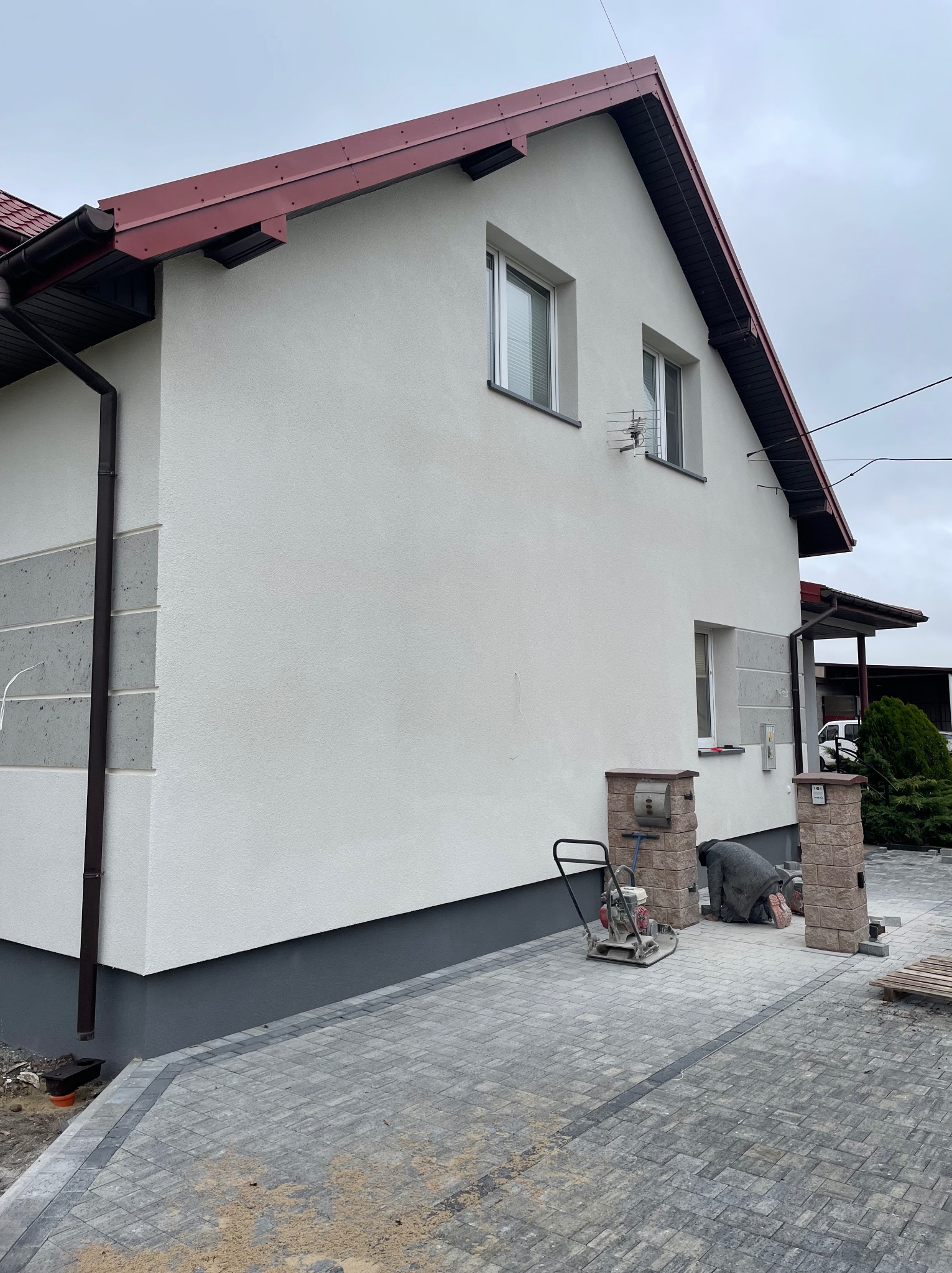 Dwukondygnacyjny dom z jasną elewacją i czerwoną dachówką. Ułożona kostka brukowa, widoczny pracownik i zagęszczarka. Detal: dekoracyjne wykończenie elewacji.