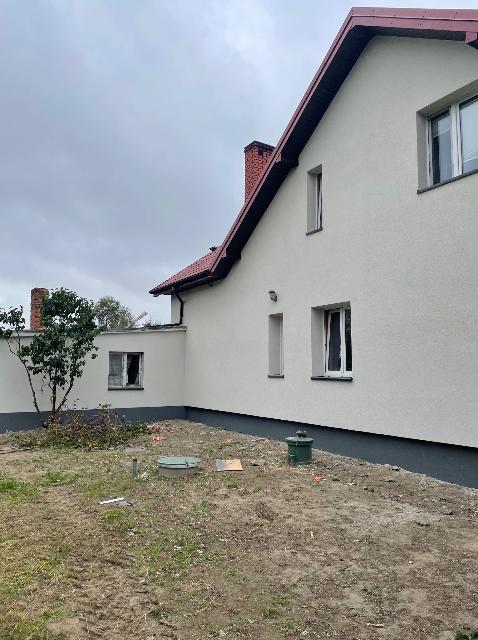 Boczny widok świeżo otynkowanego domu z czerwonym dachem i kominem. Na pierwszym planie ziemia z elementami budowlanymi i zielonymi elementami infrastruktury.