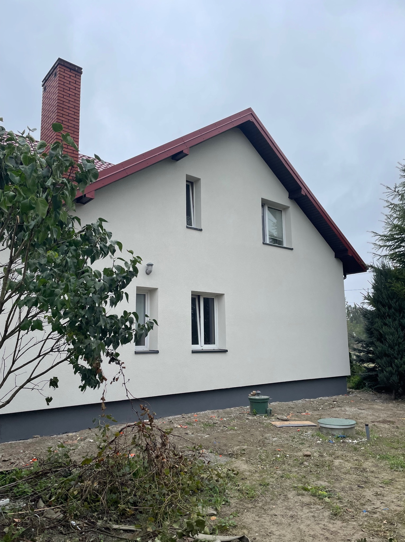 Biały dom z czerwonym dachem i ceglanym kominem, widziany z boku, z zielonym drzewem z przodu. Świeżo otynkowana elewacja i pomalowana podmurówka.