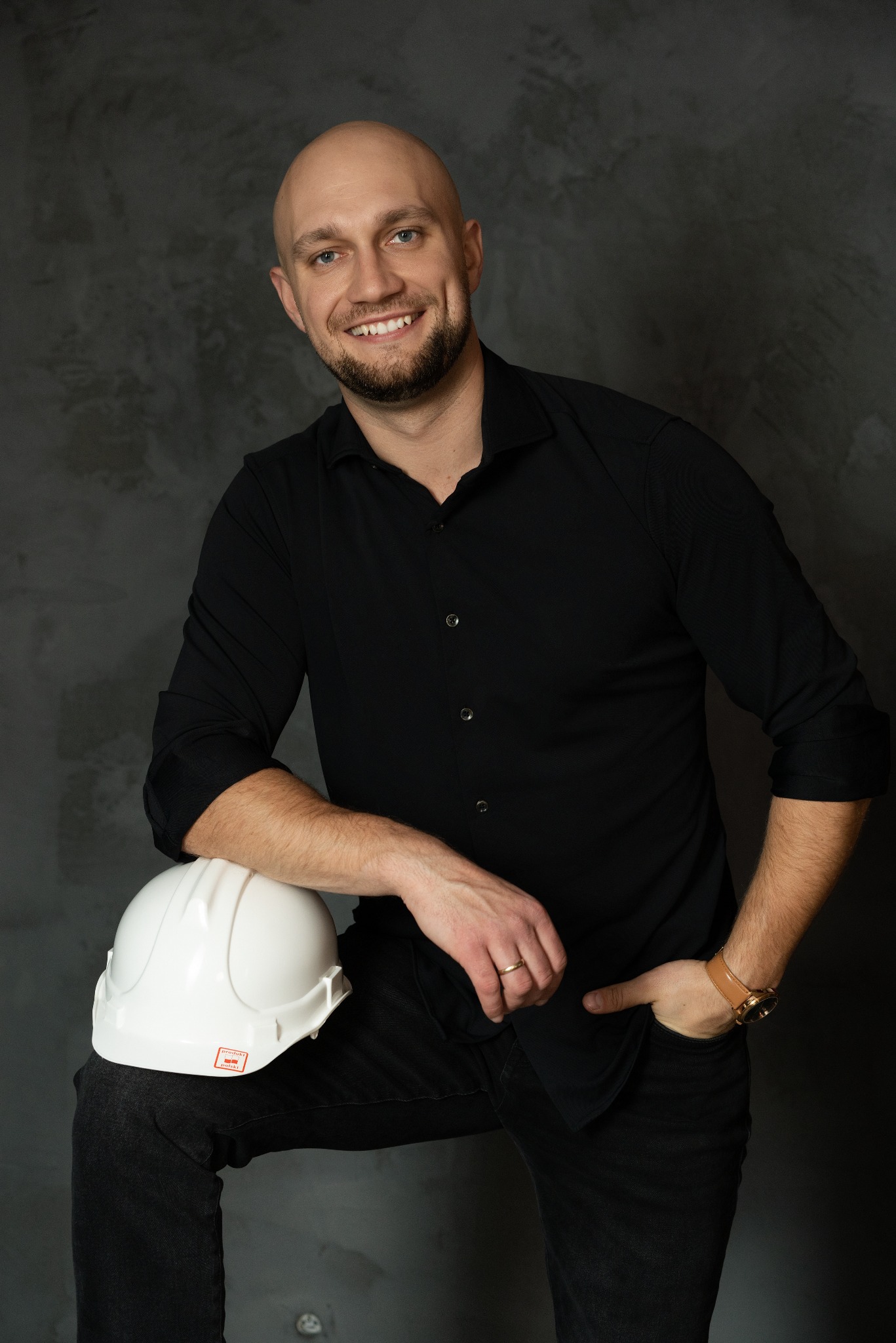 Uśmiechnięty inżynier budownictwa w czarnej koszuli, oparty o biały kask na tle szarej ściany. Widoczny zegarek i obrączka. Profesjonalny portret.