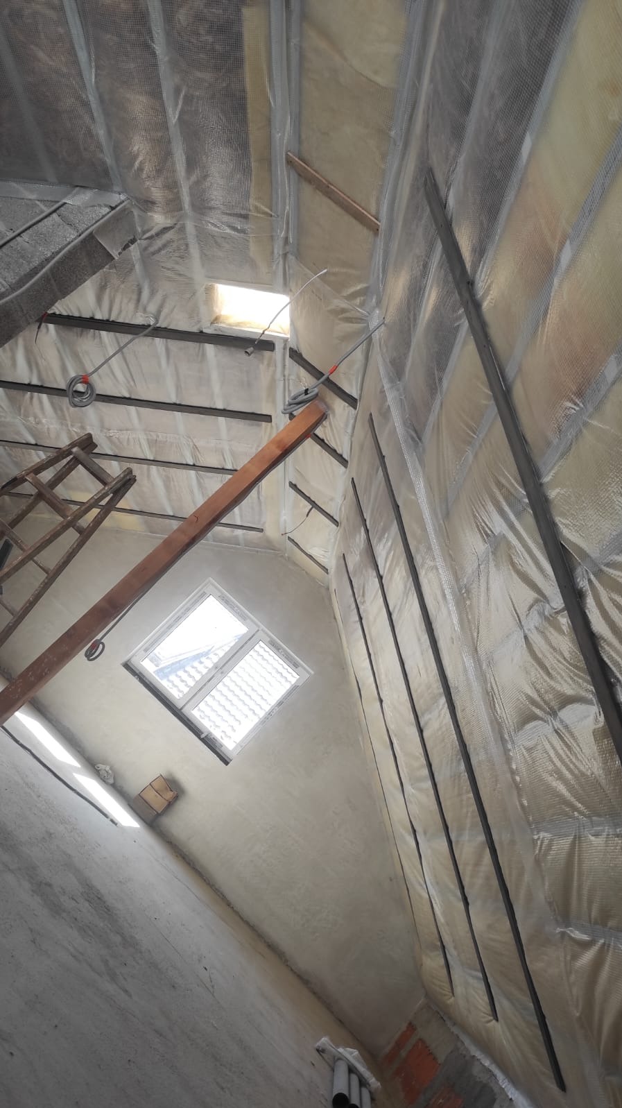 Ocieplenie poddasza z folią paroizolacyjną i metalowymi profilami, widok na okno dachowe i drewnianą konstrukcję, drabina w tle. Prace wykończeniowe.
