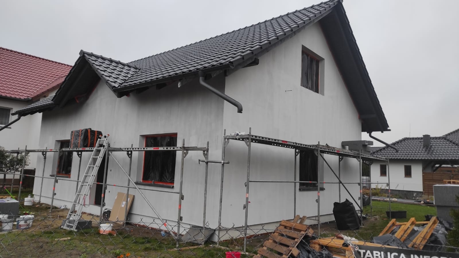Dom jednorodzinny w trakcie ocieplania elewacji, widoczne rusztowania, okna zabezpieczone folią, świeżo otynkowana ściana w kolorze białym, dach pokryty ciemną dachówką.