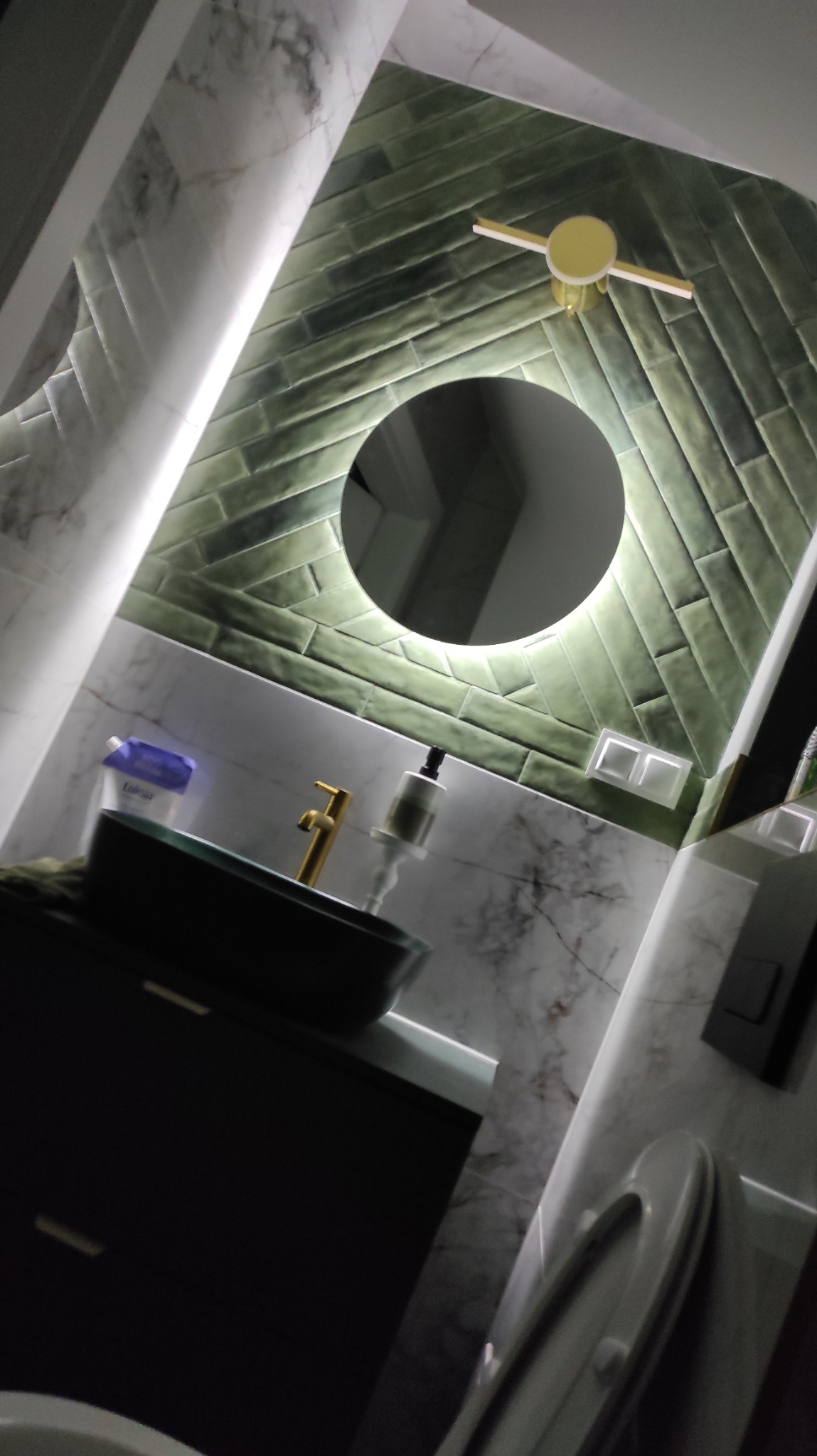 Nowoczesna łazienka z zielonymi płytkami ułożonymi w jodełkę, okrągłym lustrem z podświetleniem LED i marmurową umywalką nablatową z złotą armaturą.