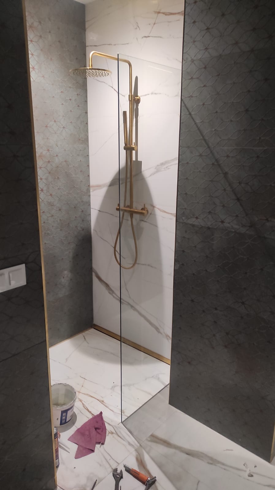 Nowoczesna kabina prysznicowa typu walk-in z złotą armaturą i marmurowymi płytkami. Widoczne narzędzia i akcesoria montażowe.