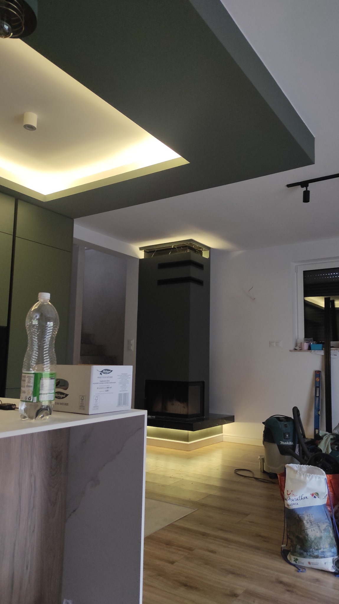 Nowoczesne wnętrze z kominkiem obudowanym szarą płytą, podświetlonym od dołu. Widoczny fragment blatu i podłogi. Sufit z oświetleniem ledowym.
