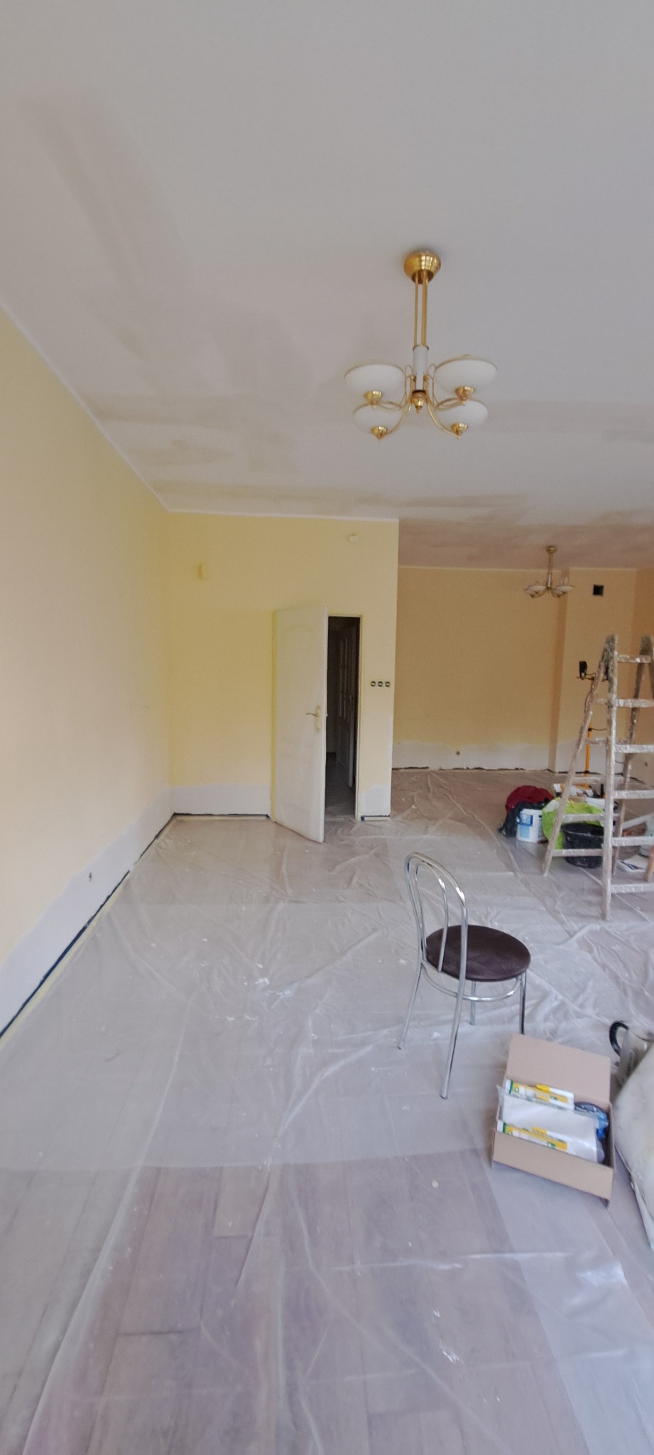 Pokój w trakcie odświeżania: żółte ściany, zabezpieczona folią podłoga, drabina malarska w tle. Widoczny otwarty karton z akcesoriami malarskimi i krzesło.