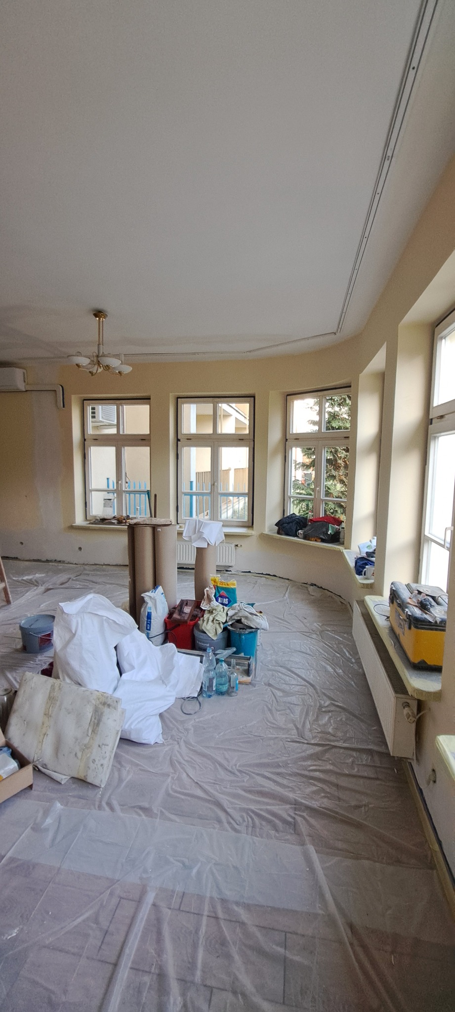 Pokój w trakcie remontu: podłoga zabezpieczona folią, widoczne farby, wiadra i narzędzia malarskie. Ściany w odcieniach beżu, trzy okna z widokiem na zieleń.