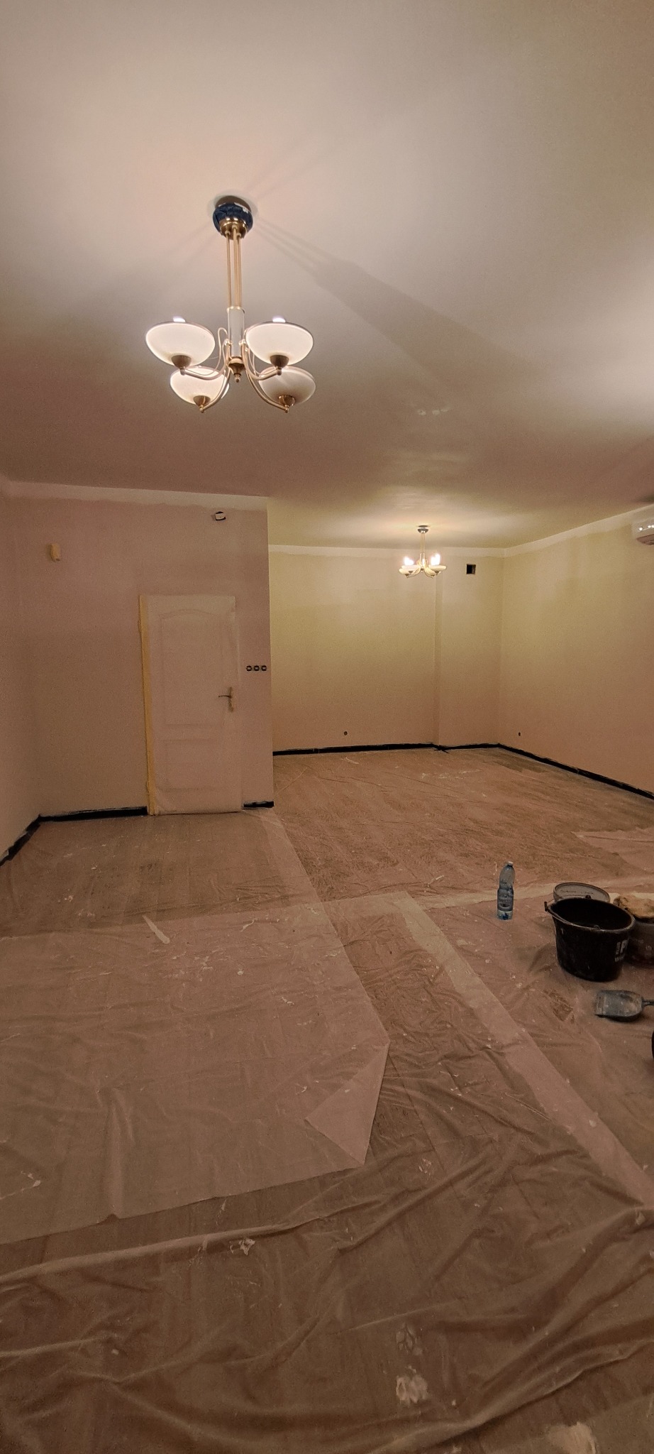 Przygotowane do malowania wnętrze z zabezpieczoną folią podłogą, widoczne narzędzia malarskie i oświetlenie sufitowe. Ściany po szpachlowaniu, gotowe do nałożenia farby.