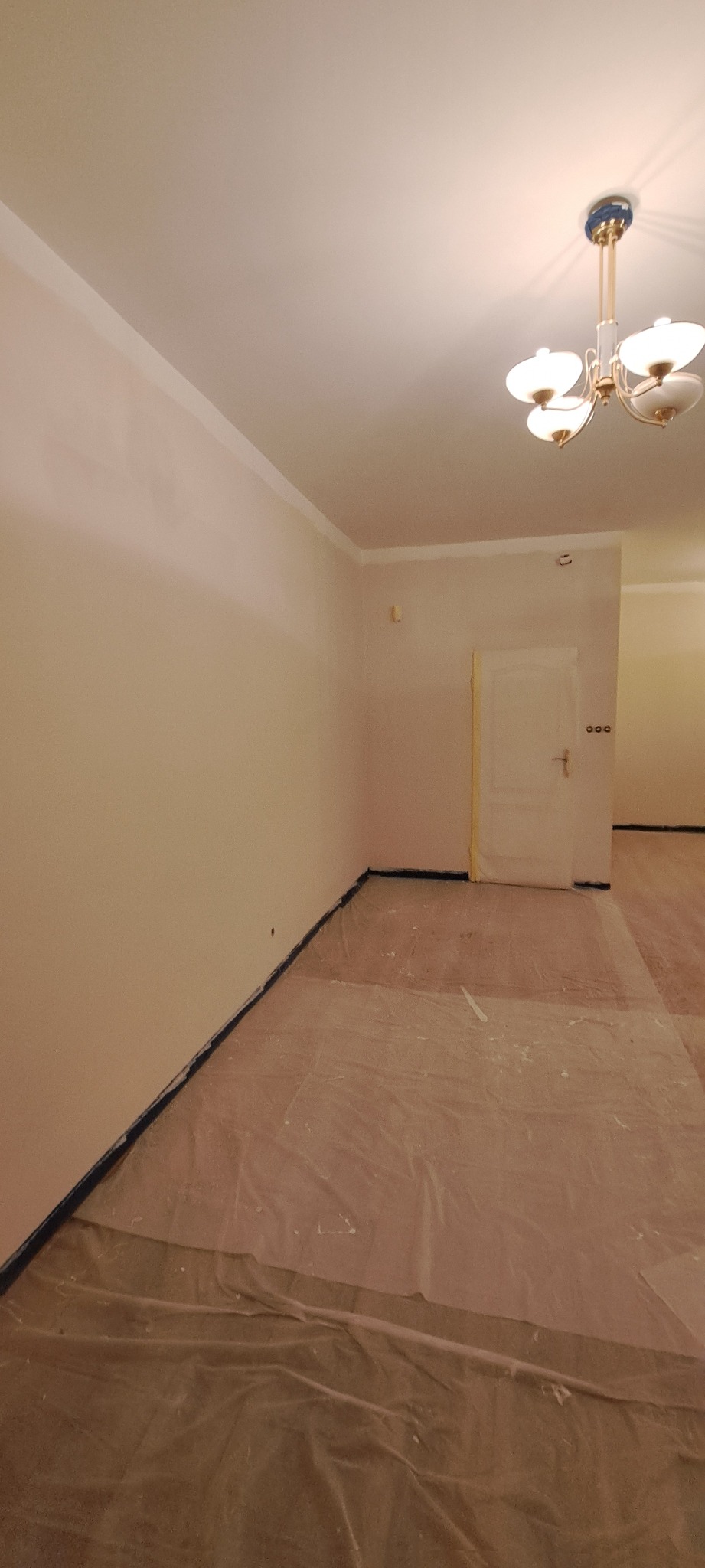 Pokój po malowaniu, zabezpieczony folią malarską. Widoczne świeżo pomalowane ściany w jasnym odcieniu, listwy przypodłogowe zabezpieczone taśmą, sufit z ozdobnym żyrandolem.