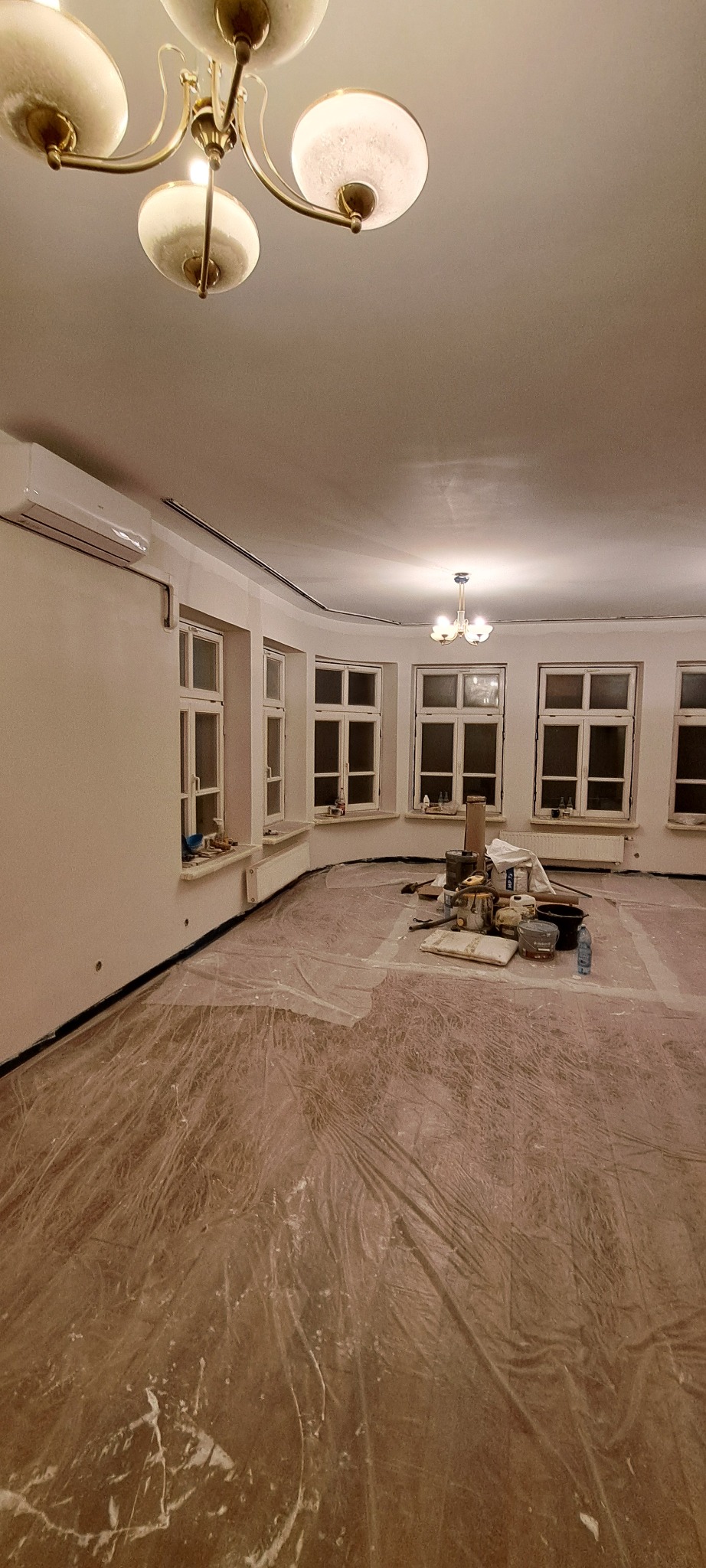 Pokój przed malowaniem: zabezpieczona folią podłoga, okna z parapetami, ściany po szpachlowaniu, widoczny klimatyzator i narzędzia malarskie.