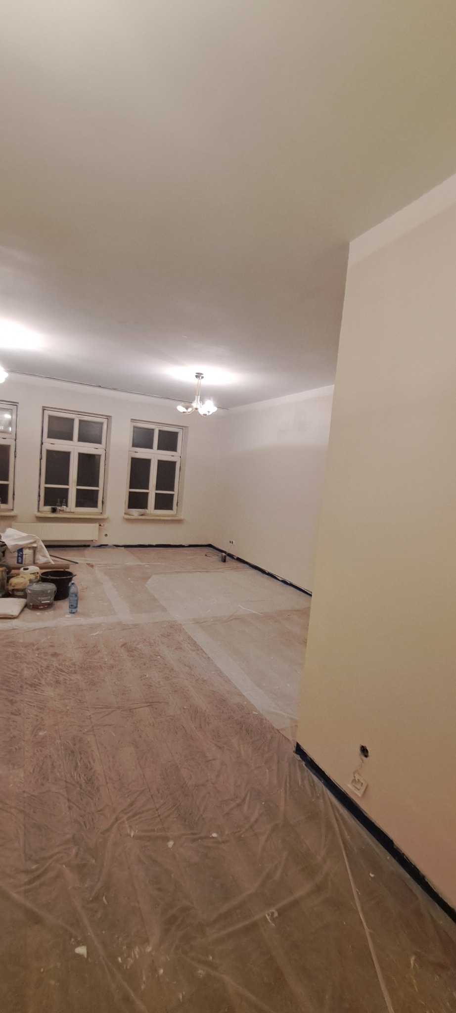 Pokój po świeżym malowaniu ścian na jasny odcień, zabezpieczona podłoga folią malarską, widoczne okna i sufit z lampą. Przygotowania do dalszych prac wykończeniowych.
