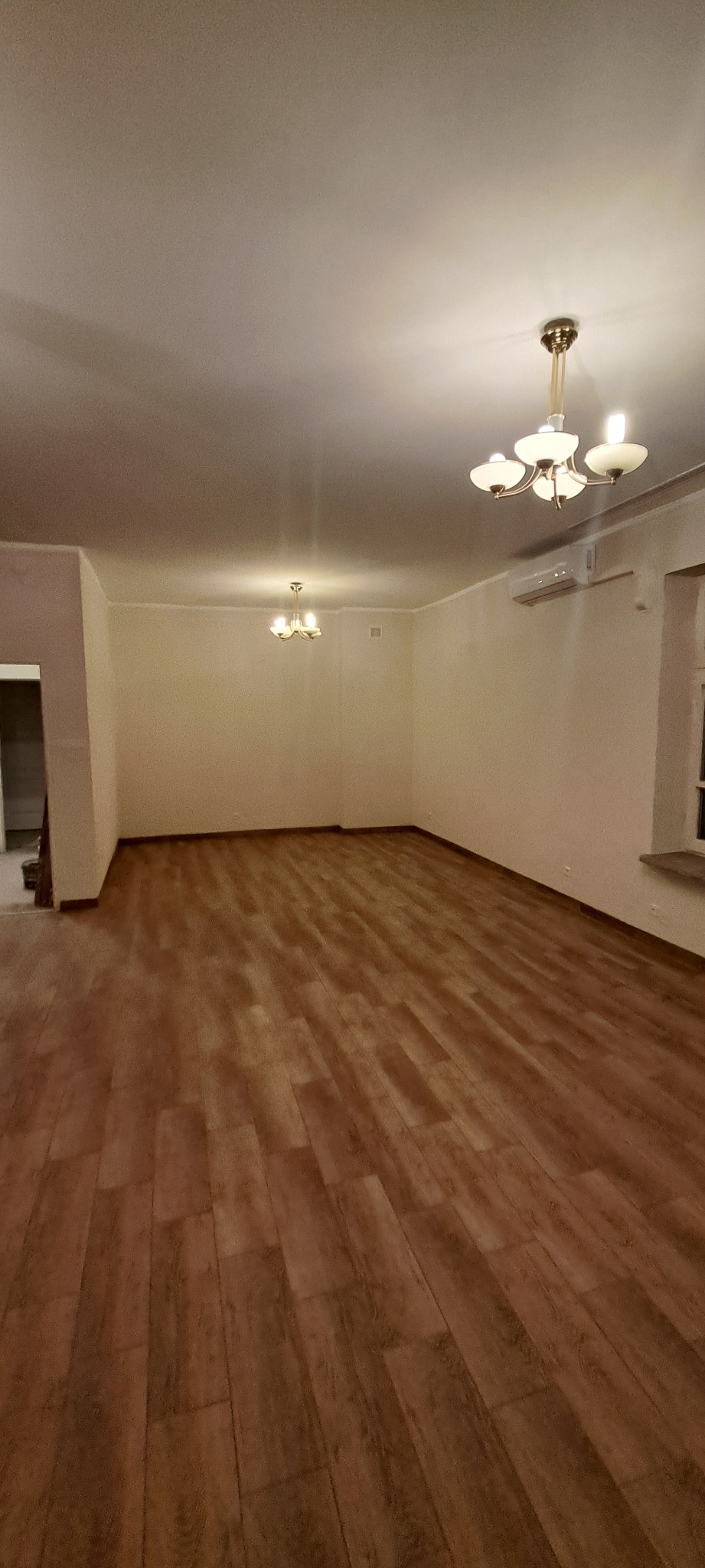 Jasne panele podłogowe w przestronnym, odnowionym pokoju z kremowymi ścianami i dwoma ozdobnymi żyrandolami. Widoczny klimatyzator.