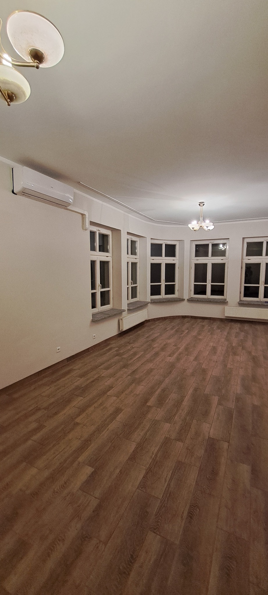 Ukończone układanie paneli podłogowych w przestronnym, jasnym pokoju z dużymi oknami. Widoczny parkiet w odcieniach brązu i beżu, białe ściany i sufit z lampą.