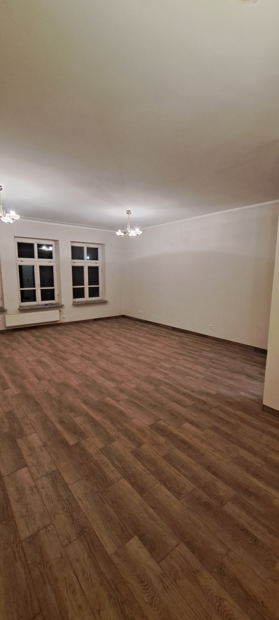 Jasne wnętrze po remoncie: brązowe panele podłogowe ułożone w jodełkę, białe ściany i okna. Widoczne lampy sufitowe i grzejnik pod oknem.