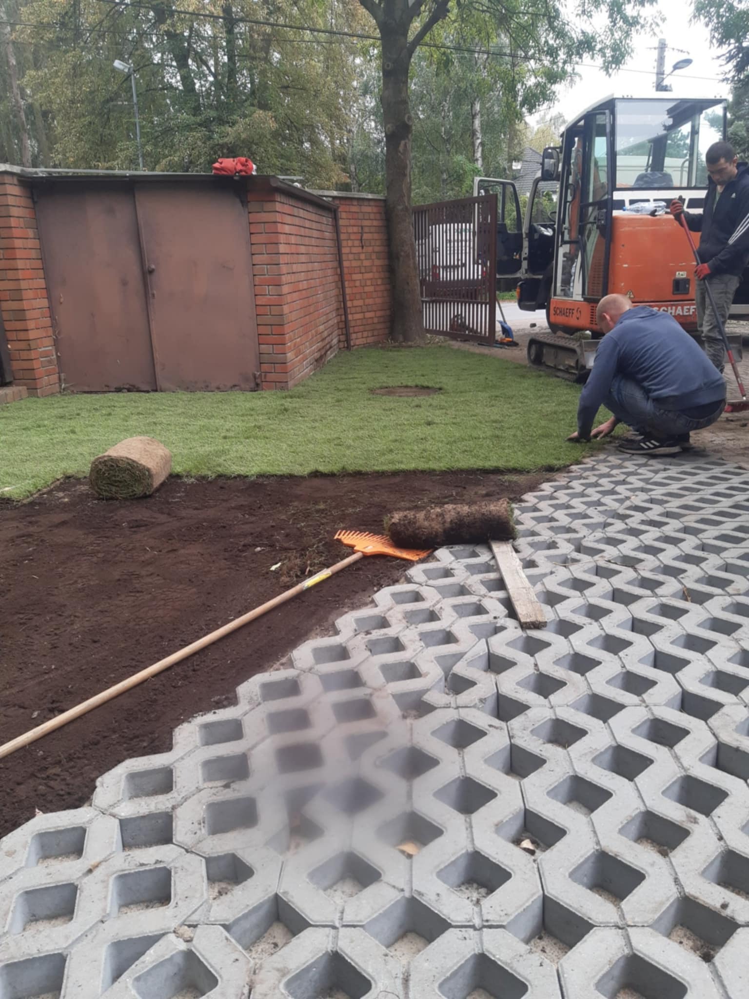 Układanie trawy z rolki i ażurowych płyt betonowych na posesji. W tle widoczny minikoparka i pracownicy. Częściowo ułożona trawa i płyty.