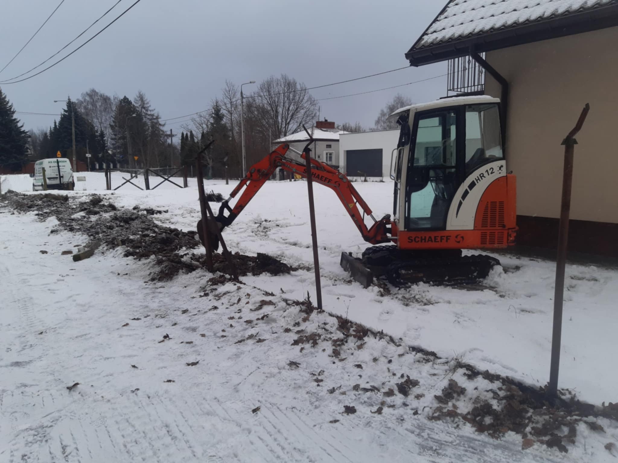Pomarańczowa mini koparka Schaeff HR12 w zimowym krajobrazie, wykonująca wykop w zasypanym śniegiem terenie. W tle budynek i drzewa.