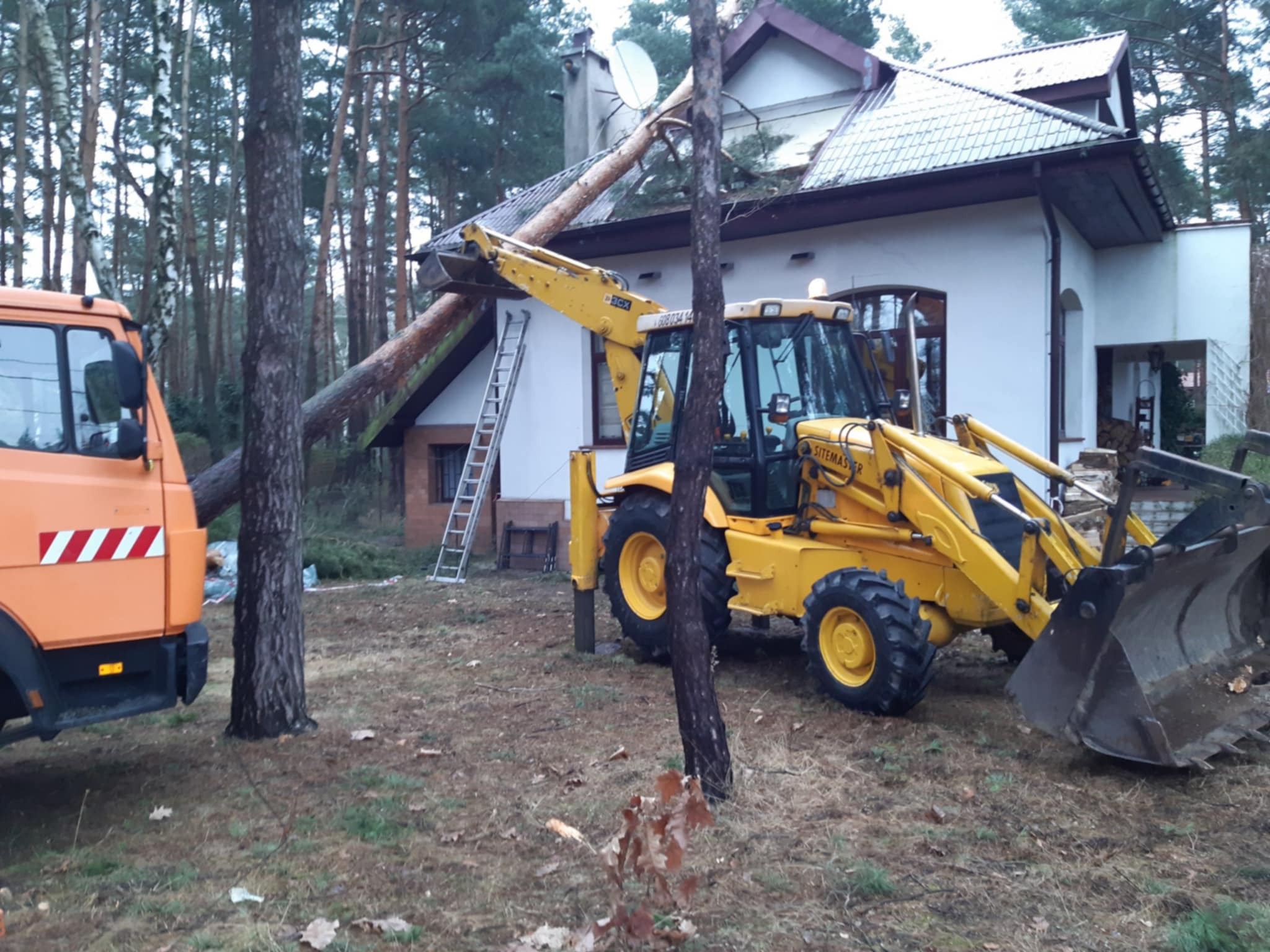 Żółta koparko-ładowarka usuwa powalone drzewo z dachu białego domu. W tle las i pomarańczowa ciężarówka. Drabina oparta o ścianę. Prace po wichurze.