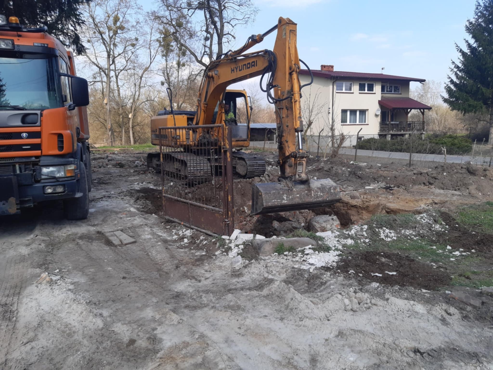 Żółta koparka Hyundai w trakcie prac ziemnych na posesji z domem w tle. Widoczny wykop i pomarańczowa ciężarówka. Prace ziemne w toku.