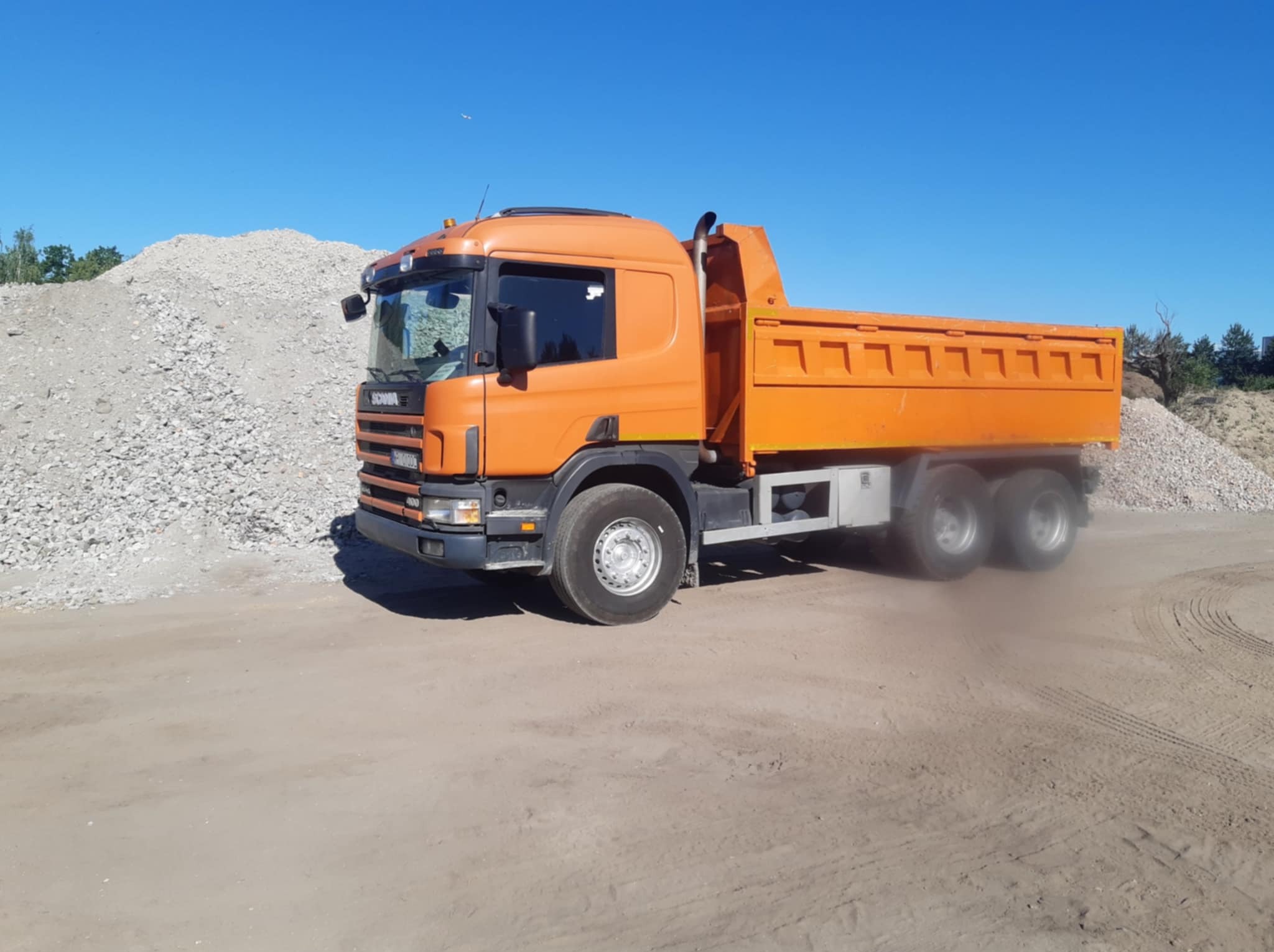 Pomarańczowa wywrotka Scania na placu budowy, z hałdą gruzu w tle, pod błękitnym niebem. Widoczne ślady opon na ziemi.