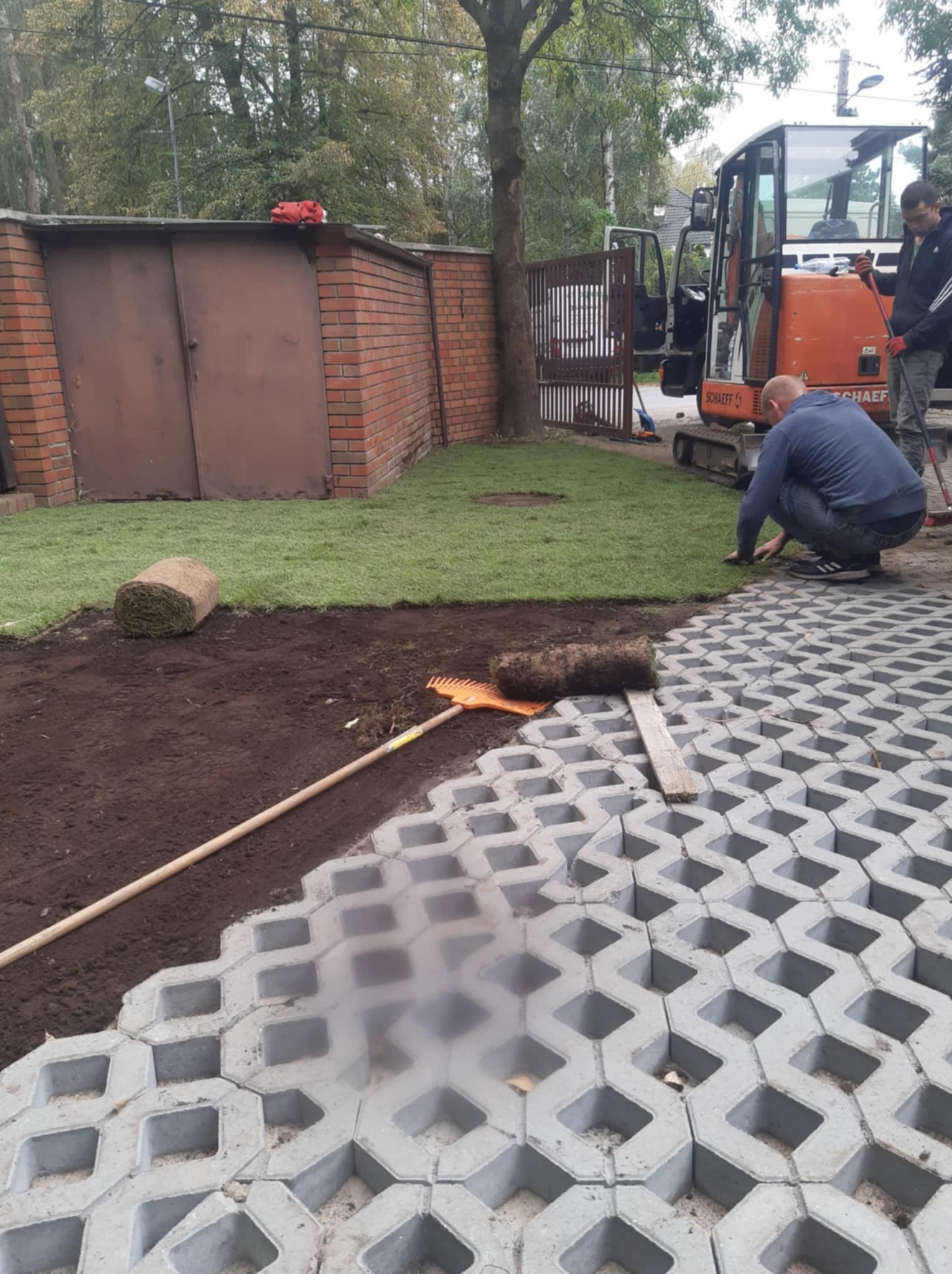 Układanie trawy z rolki i betonowej ażurowej nawierzchni na posesji. W tle mini koparka i pracownicy. Widoczny etap prac z wykorzystaniem narzędzi.