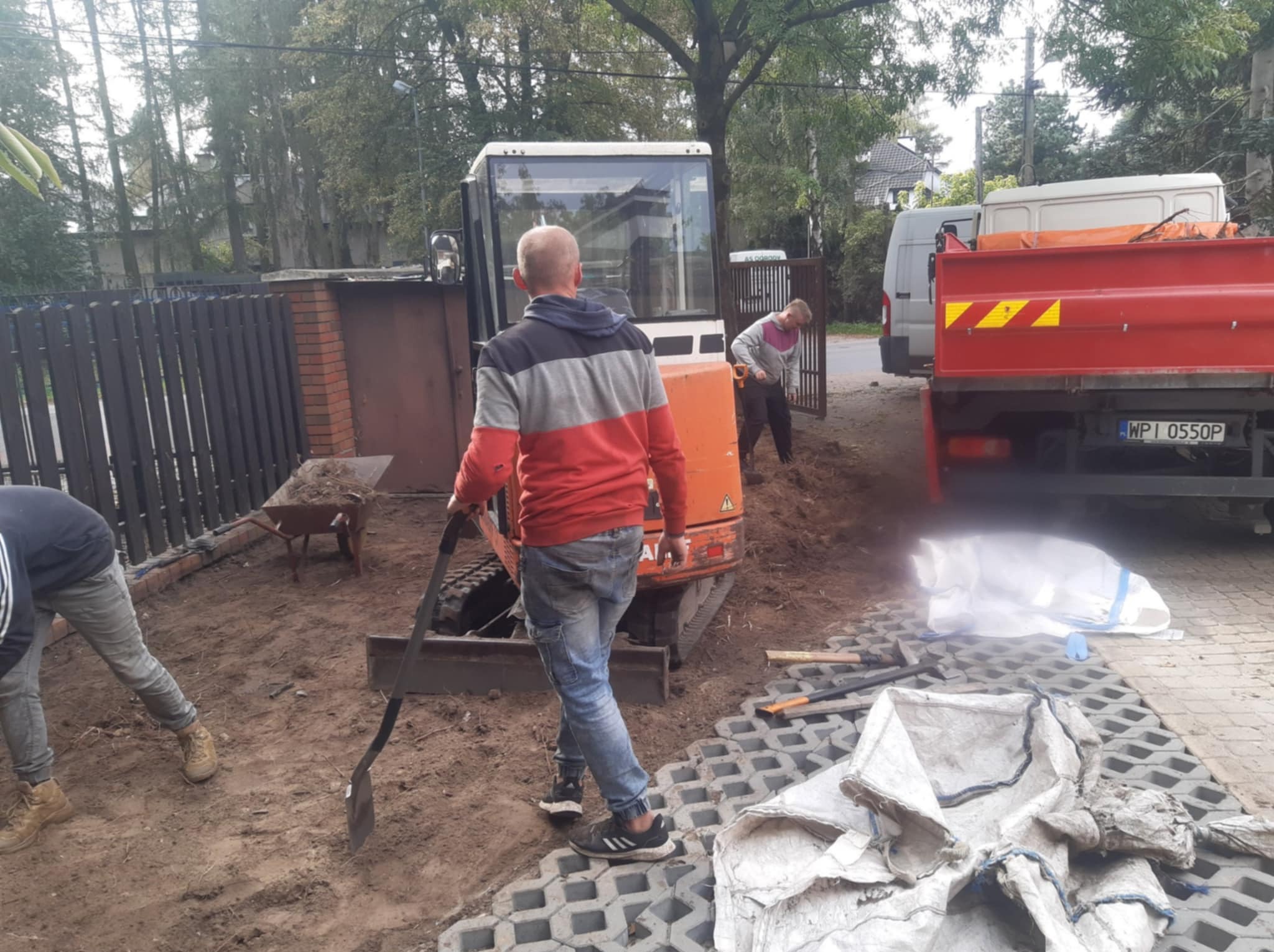 Prace ziemne: mini koparka, pracownicy z łopatami i wywrotka na posesji. Przygotowanie terenu pod budowę, widoczne eko-kratki i narzędzia.