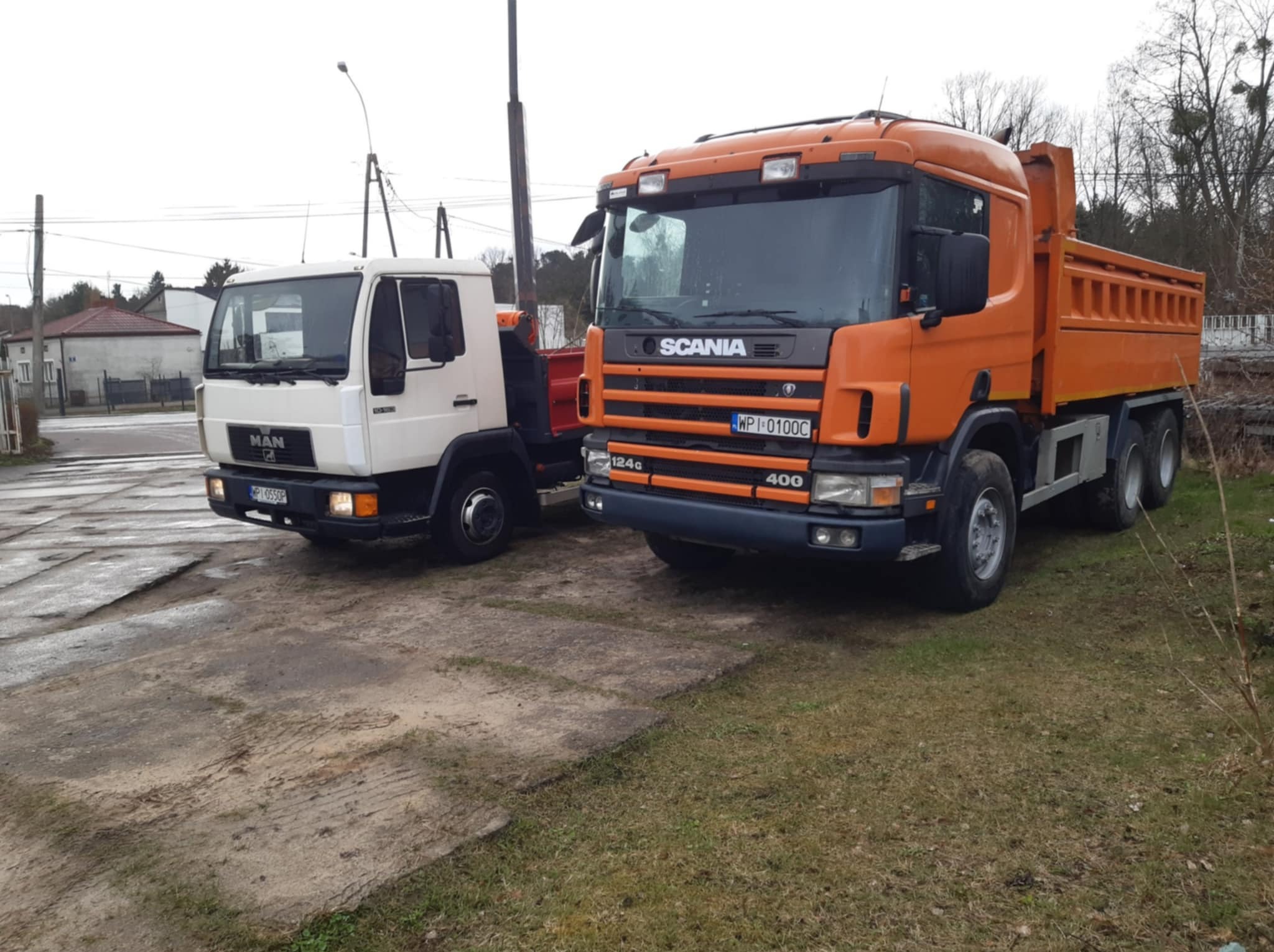 Dwa samochody ciężarowe: biały MAN i pomarańczowy Scania 124c 400, stojące na placu. Widoczne tablice rejestracyjne. Tło stanowią budynki i drzewa.