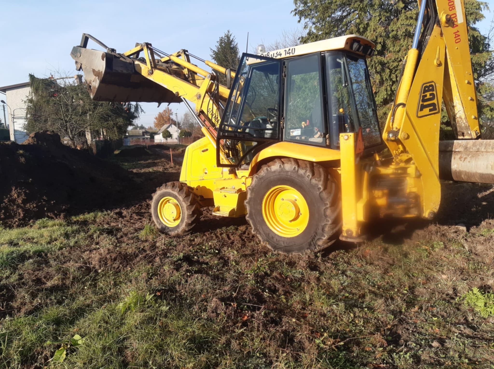 Żółta koparko-ładowarka JCB na placu budowy, z wykopaną ziemią w tle. Widoczne detale maszyny i otoczenie, w tym budynki w oddali i drzewa.