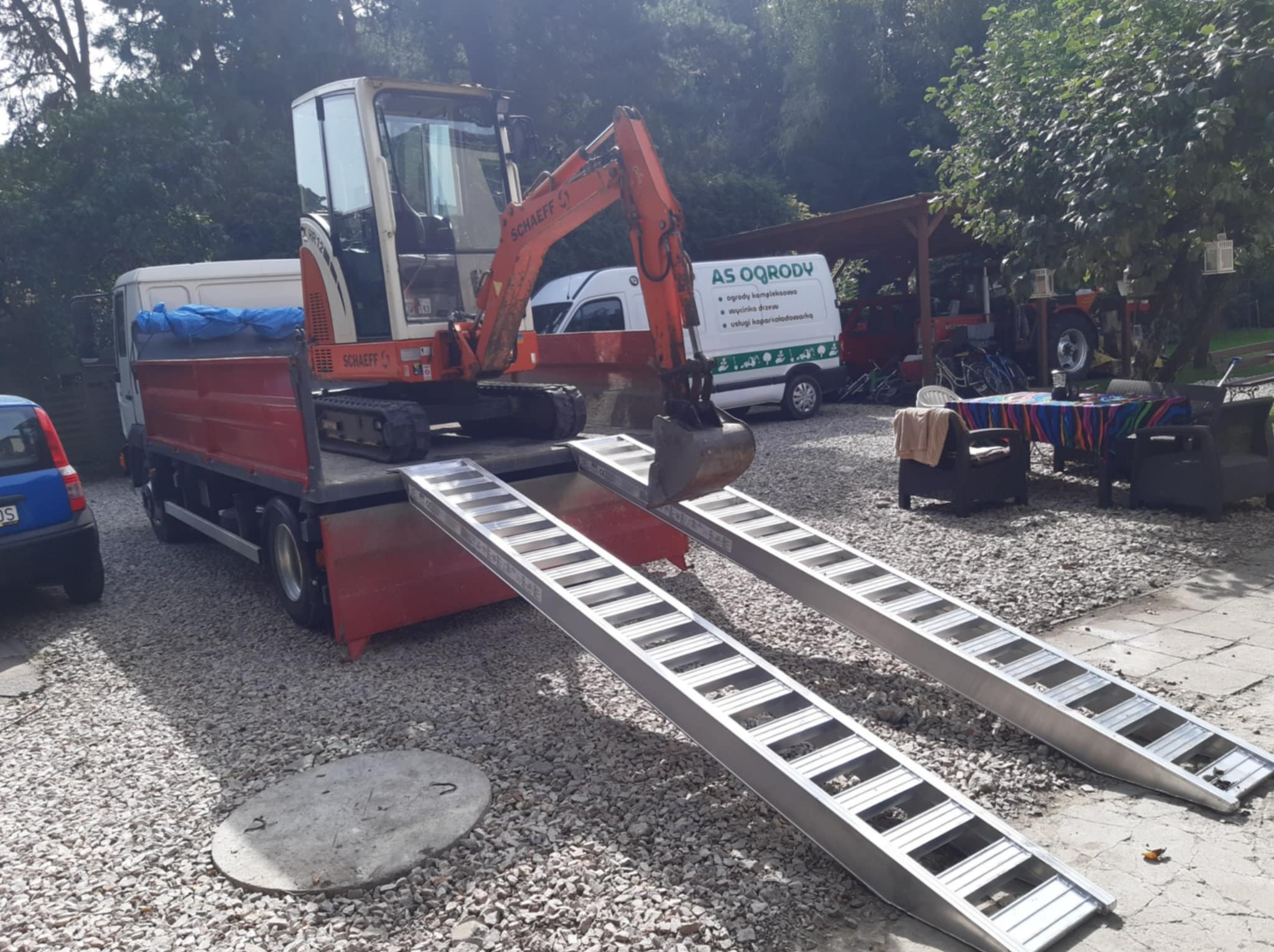 Pomarańczowa mini koparka Schaeff załadowana na czerwoną ciężarówkę z rampami na żwirowym podłożu, w tle zieleń i biały bus z logo firmy.