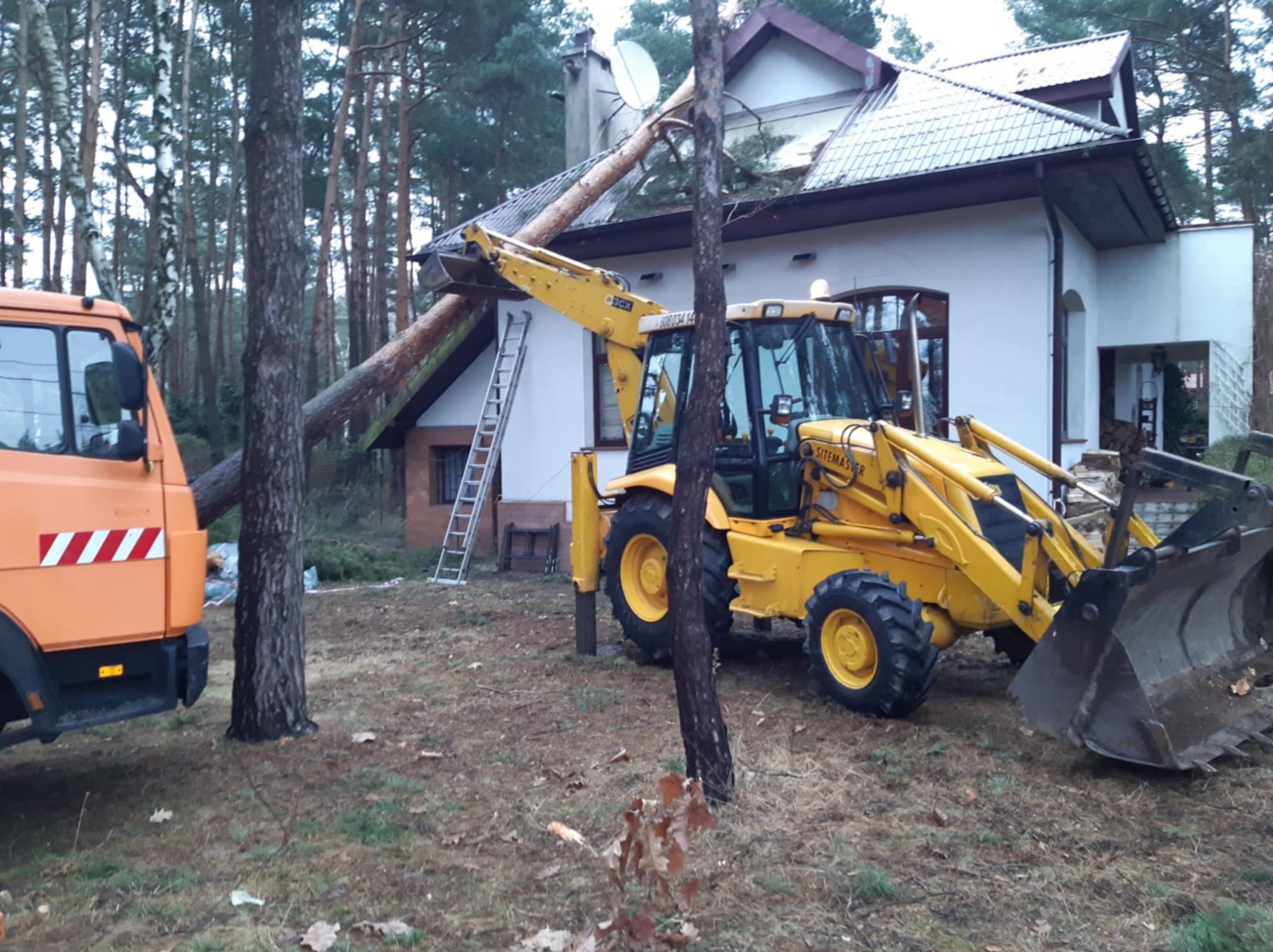 Żółta koparko-ładowarka podtrzymuje powalone drzewo na tle domu. Obok stoi pomarańczowa ciężarówka. Drabina oparta o ścianę budynku. Prace porządkowe po wichurze.