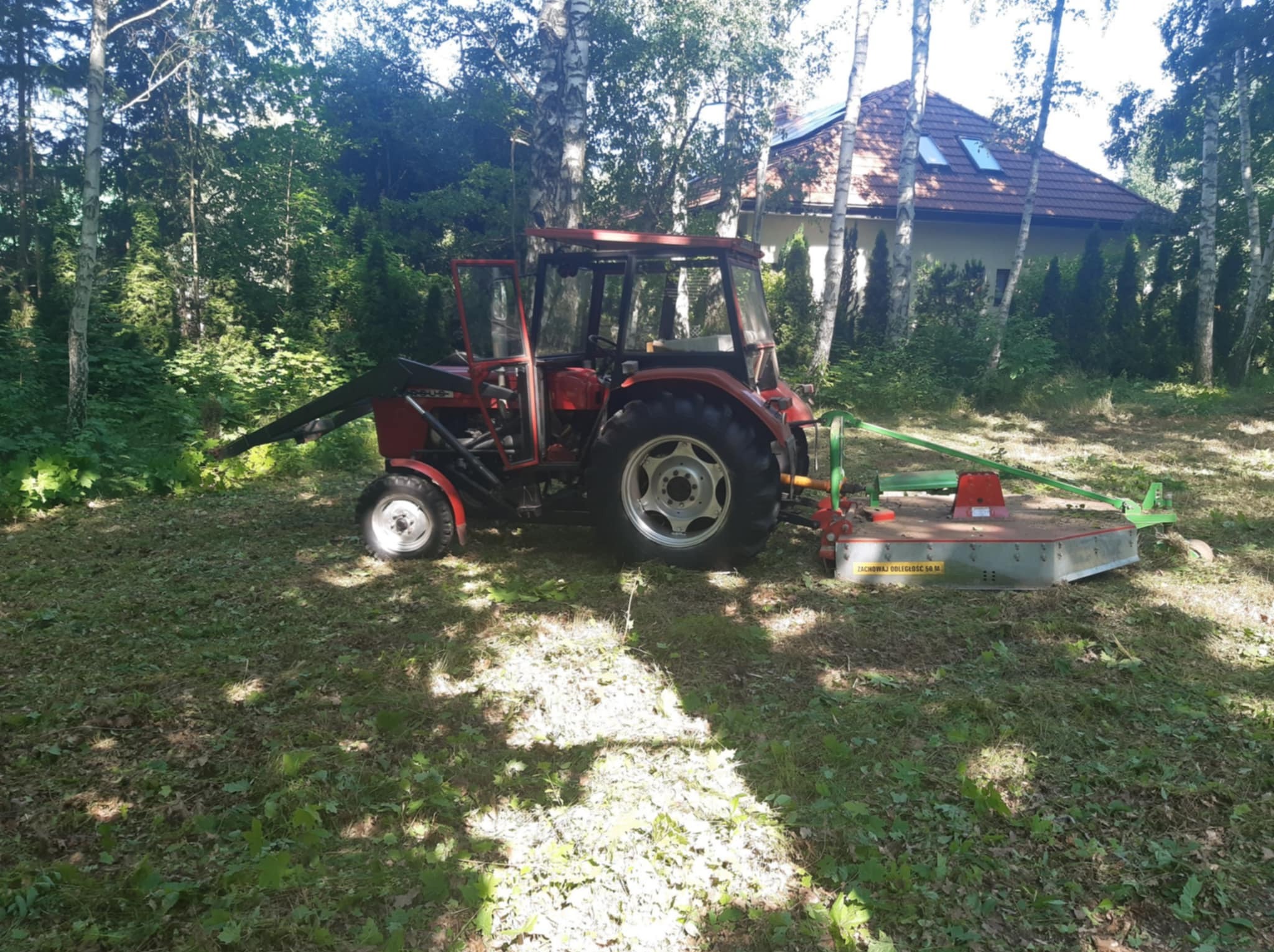 Czerwony traktor z kosiarką rotacyjną na zarośniętej działce, w tle dom jednorodzinny i drzewa. Widoczne świeżo skoszone fragmenty trawy.