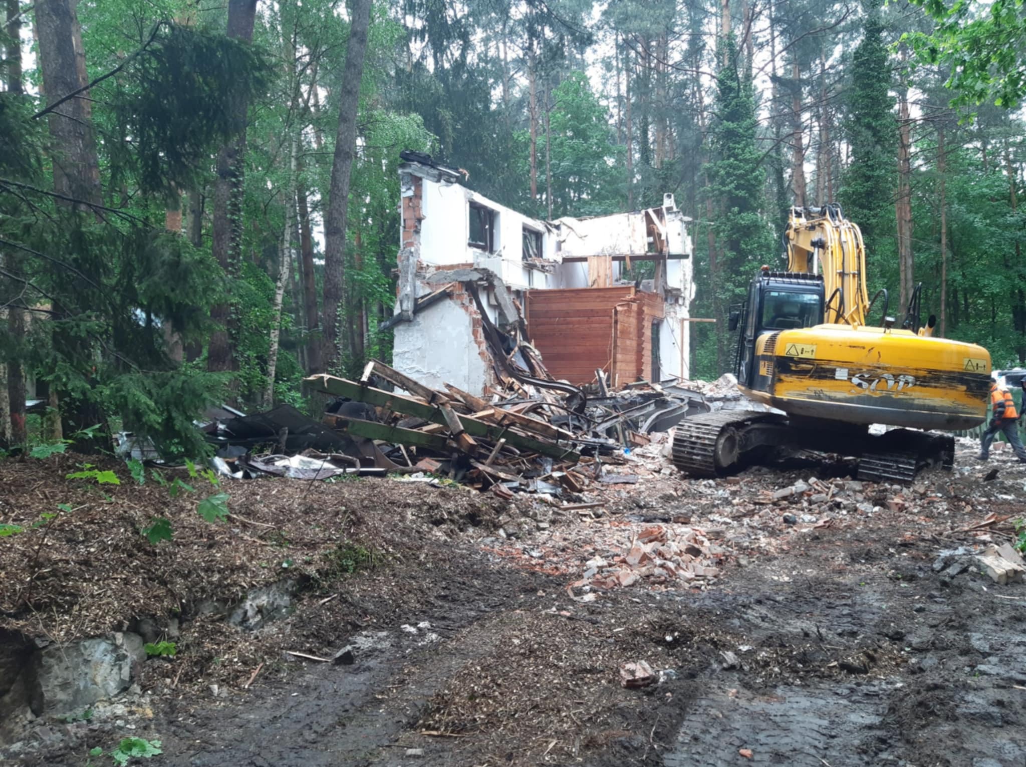Rozbiórka domu w lesie z użyciem żółtej koparki. Fragmenty ścian, gruz i drewno na ziemi. Widoczny pracownik w odblaskowej kamizelce.