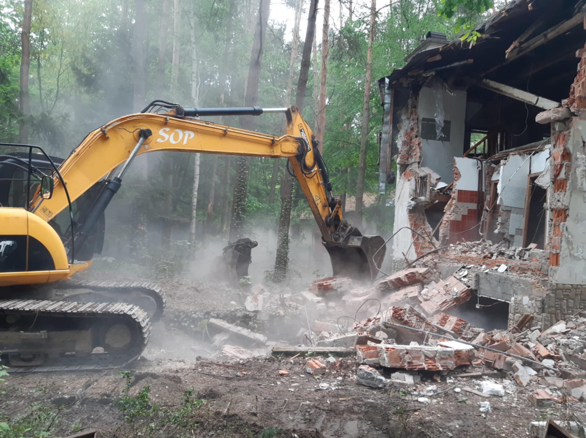 Żółta koparka SOP w akcji rozbiórki murowanego budynku w otoczeniu drzew. Gruz i pył unoszą się w powietrzu, ukazując postęp prac rozbiórkowych.