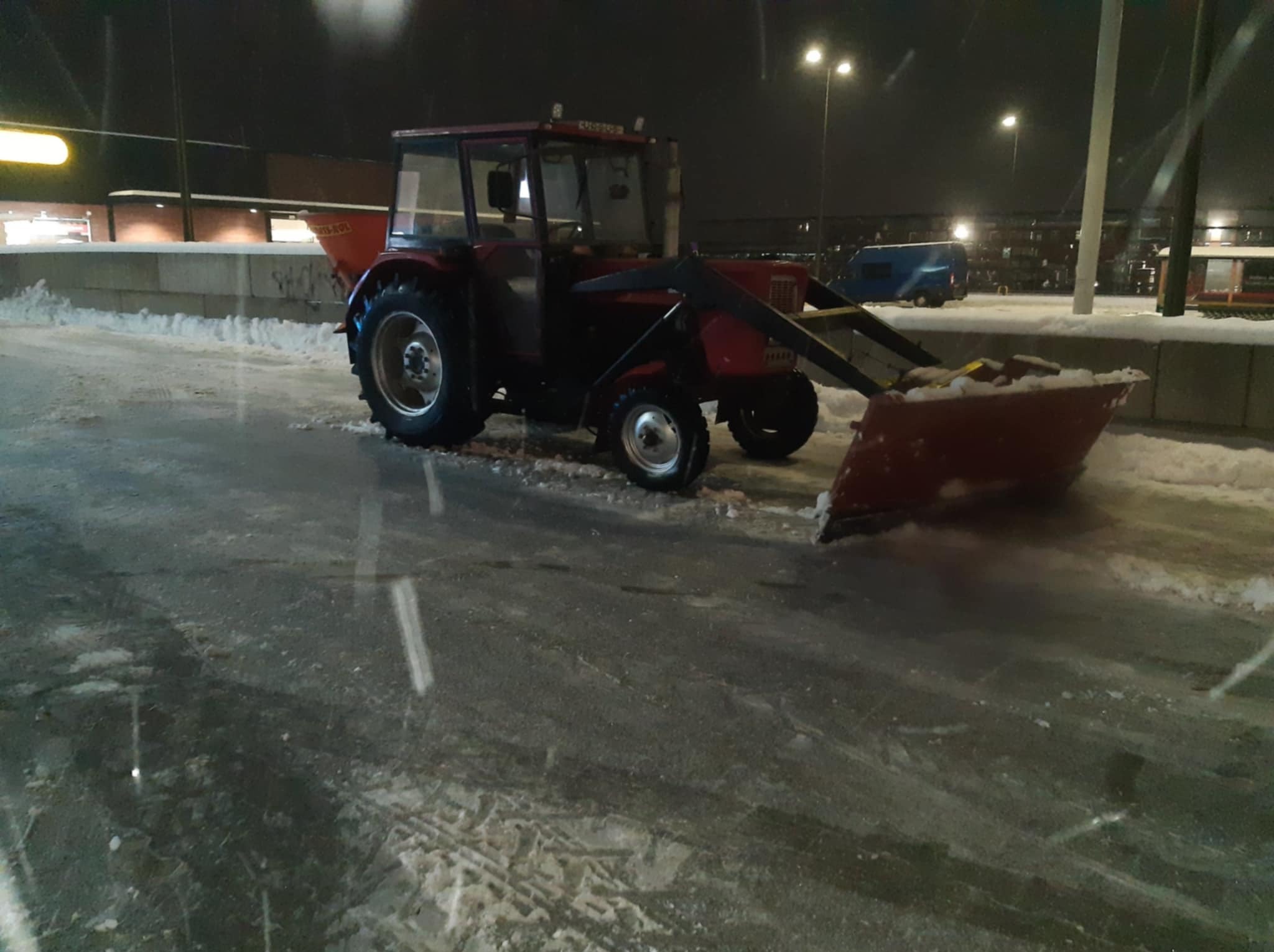 Czerwony traktor z pługiem odśnieżającym na ośnieżonej powierzchni nocą. Widoczne budynki w tle i oświetlenie uliczne. Ślady opon na śniegu.