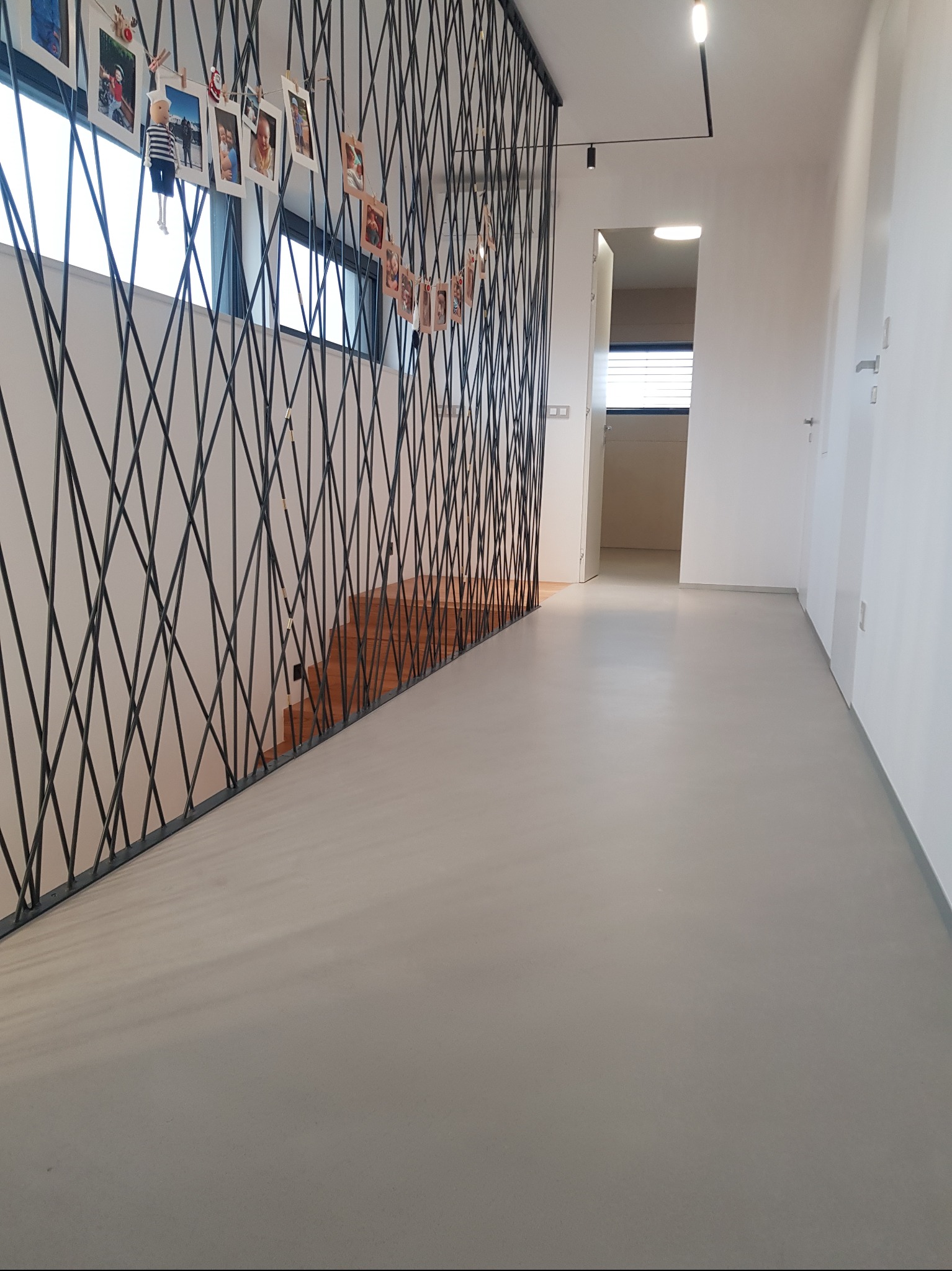 Jasnoszara posadzka w korytarzu z dekoracyjną, metalową balustradą i schodami w tle. Minimalistyczne wnętrze z białymi ścianami i oświetleniem punktowym.