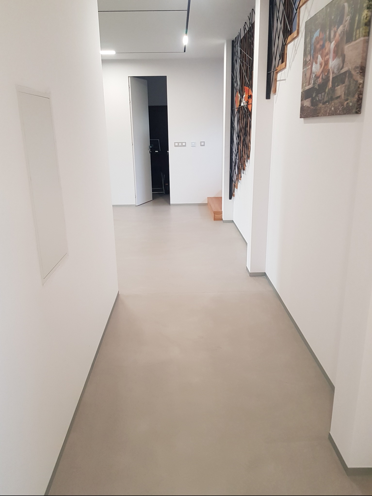 Beżowa, gładka posadzka w korytarzu z białymi ścianami i schodami z czarną balustradą. Minimalistyczny design, neutralna kolorystyka, widok na otwarte drzwi.