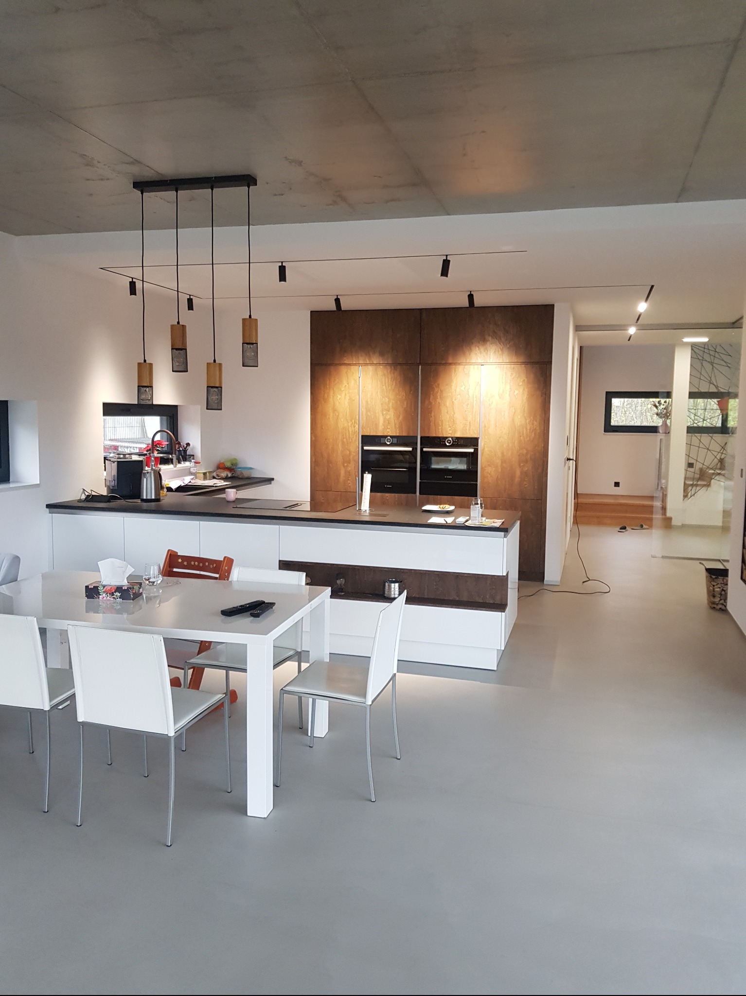 Nowoczesna kuchnia z jadalnią: szara posadzka, białe meble, drewniane akcenty. Widoczne oświetlenie punktowe i wiszące lampy. Minimalistyczny design, jasne wnętrze.