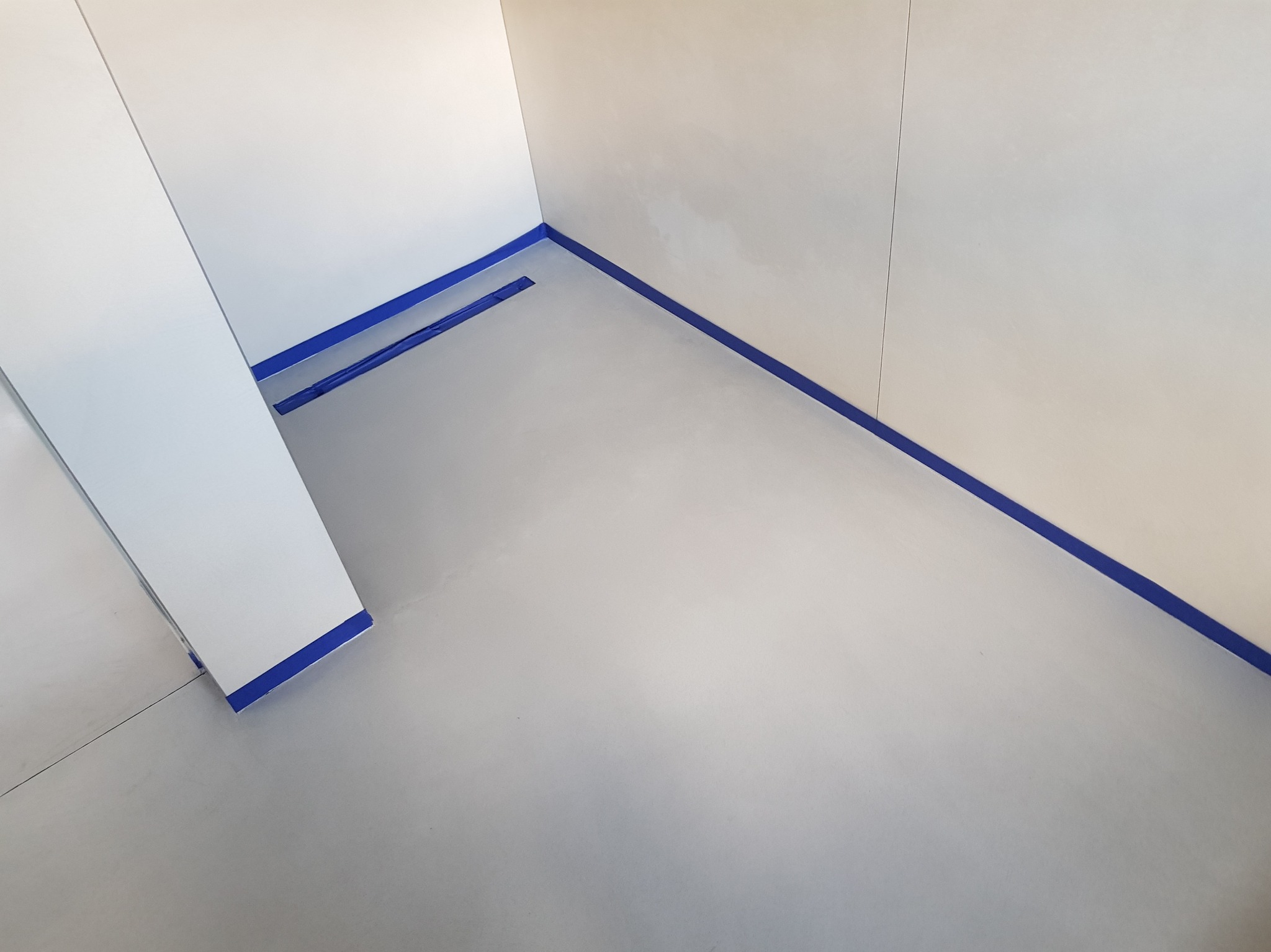 Gładka, jasnoszara posadzka w narożniku pomieszczenia zabezpieczona niebieską taśmą malarską w Wieliczce. Widoczny odpływ liniowy w podłodze.