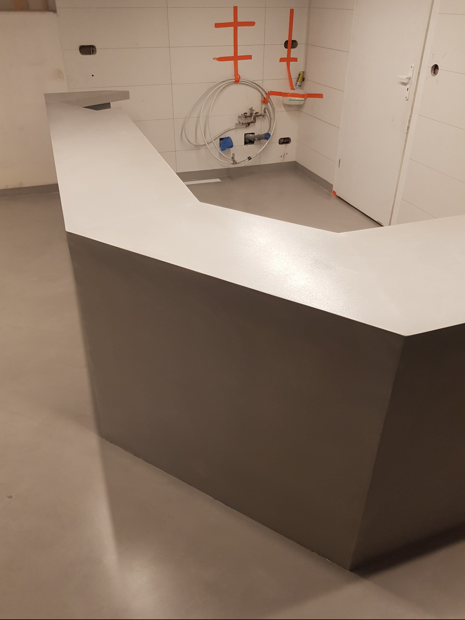 Nowoczesna łazienka z szarą posadzką i blatem w kształcie litery L, białe płytki na ścianach z widoczną instalacją wodną zabezpieczoną pomarańczową taśmą.